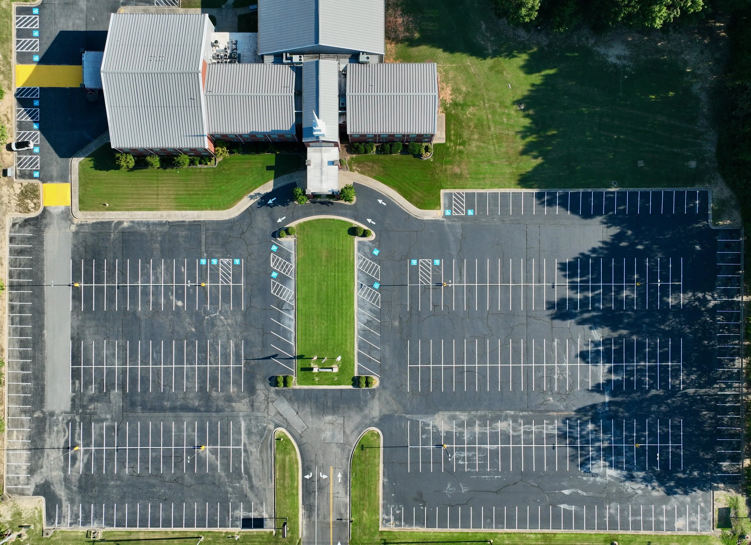 An aerial view of a mostly empty parking lot with a small grassy area and a building at the top center. The parking lot has multiple designated handicapped parking spots and clear white lines marking spaces. Shadows of trees are cast over the parking
