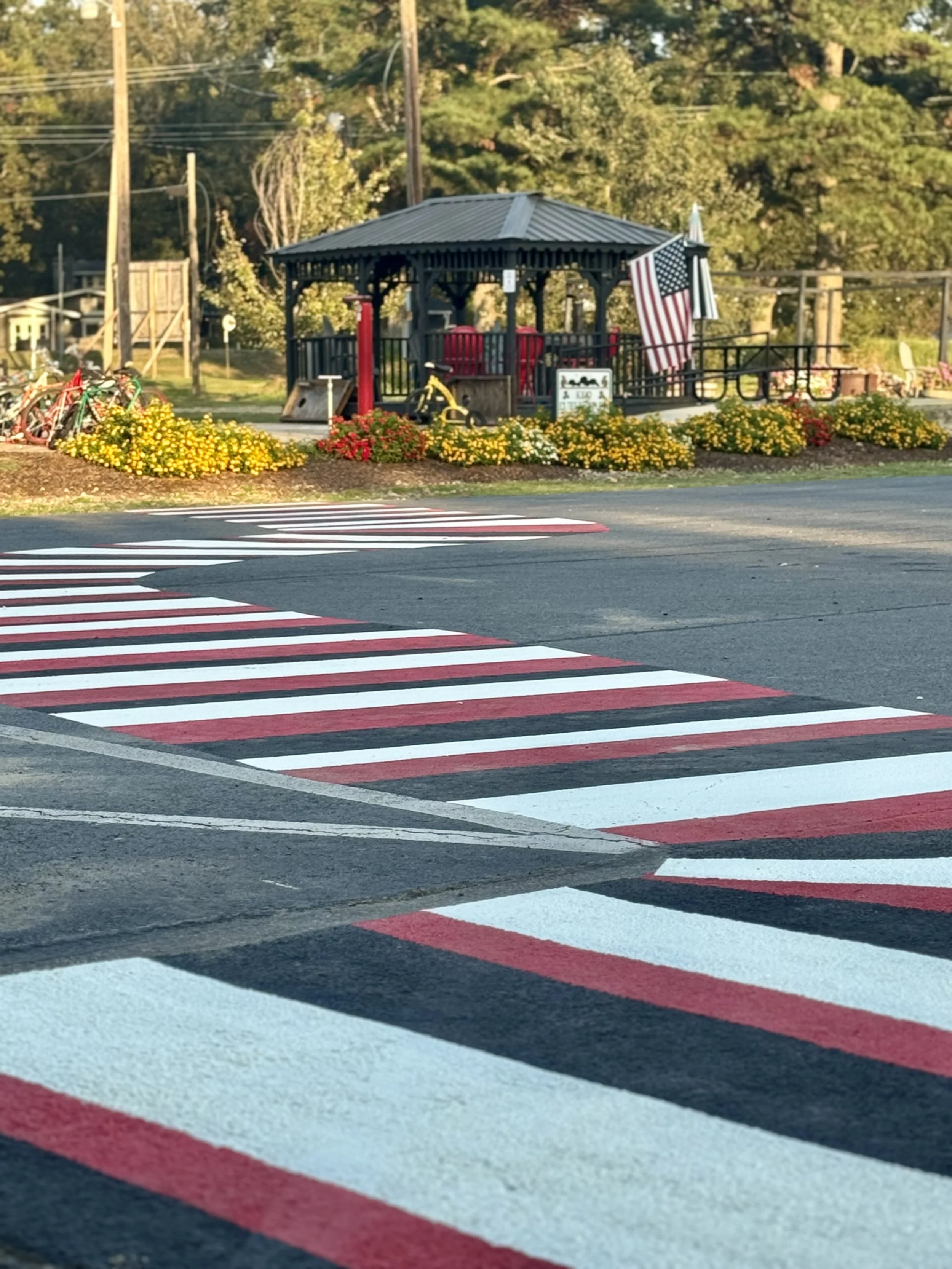 A pedestrian crosswalk with red, white, and black stripes in the foreground leads to a park with a gazebo, American flags, yellow flowers, and bicycles in the background.