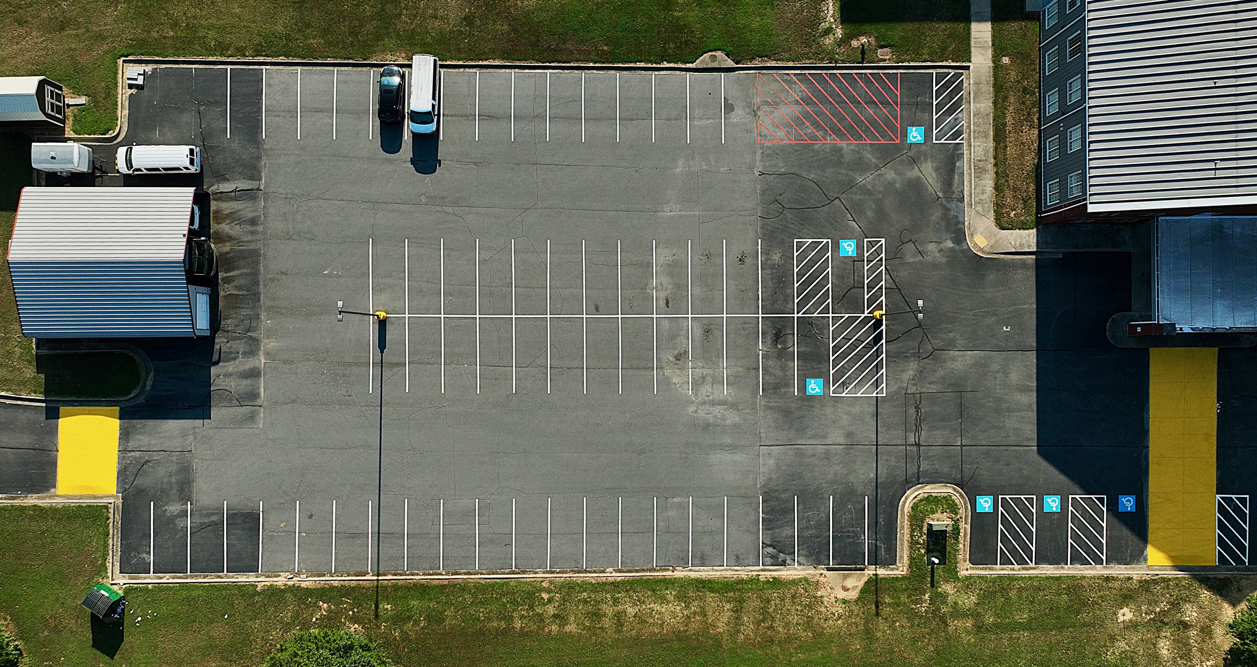 Aerial view of a mostly empty parking lot with marked parking spaces, including designated handicapped spots, surrounding a blue building on one side and green grass on the other.