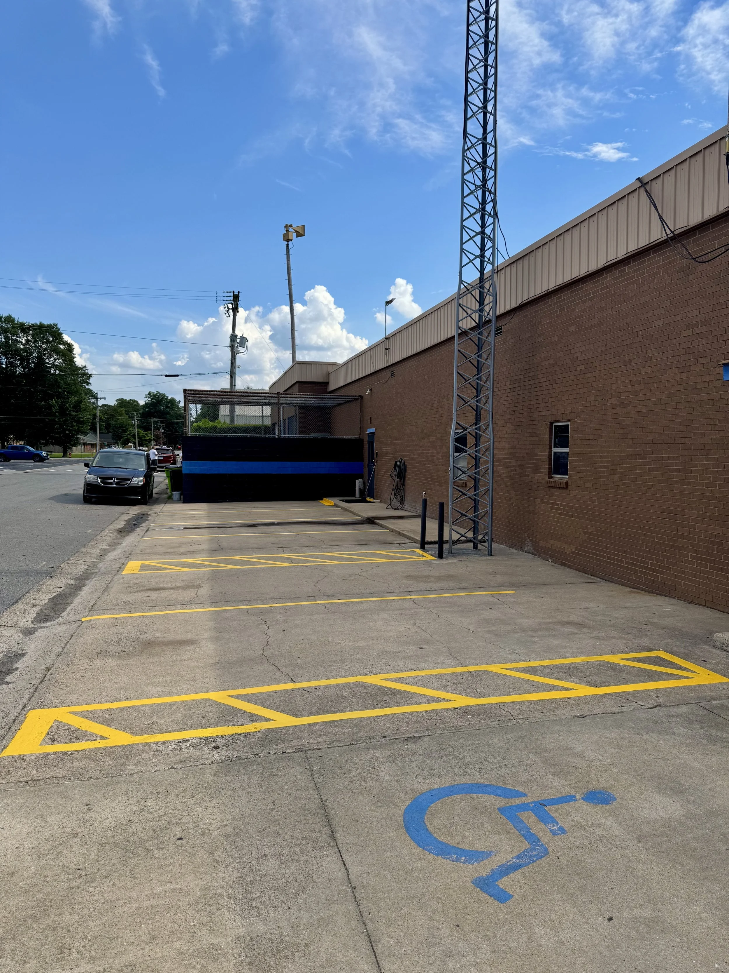 Empty parking lot with four reserved handicapped spaces marked by yellow lines, one of which is a blue handicapped symbol, next to a brick building and a street with cars, under a blue sky with some clouds.