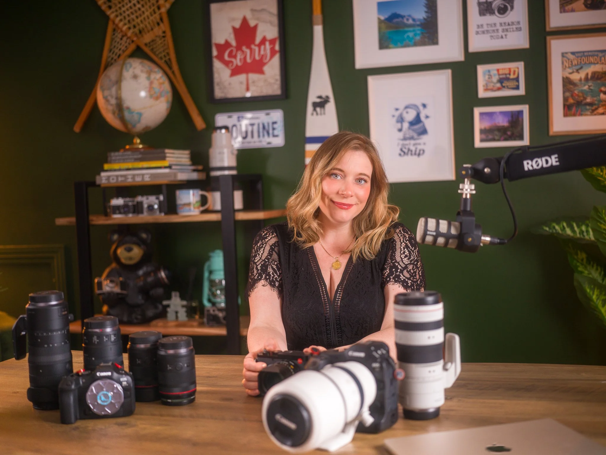 A woman sitting at a wooden table with camera equipment, looking at the camera and smiling. The table has several camera lenses and a camera. There is a microphone and recording equipment on the right side, a green wall with framed pictures and a globe in the background.