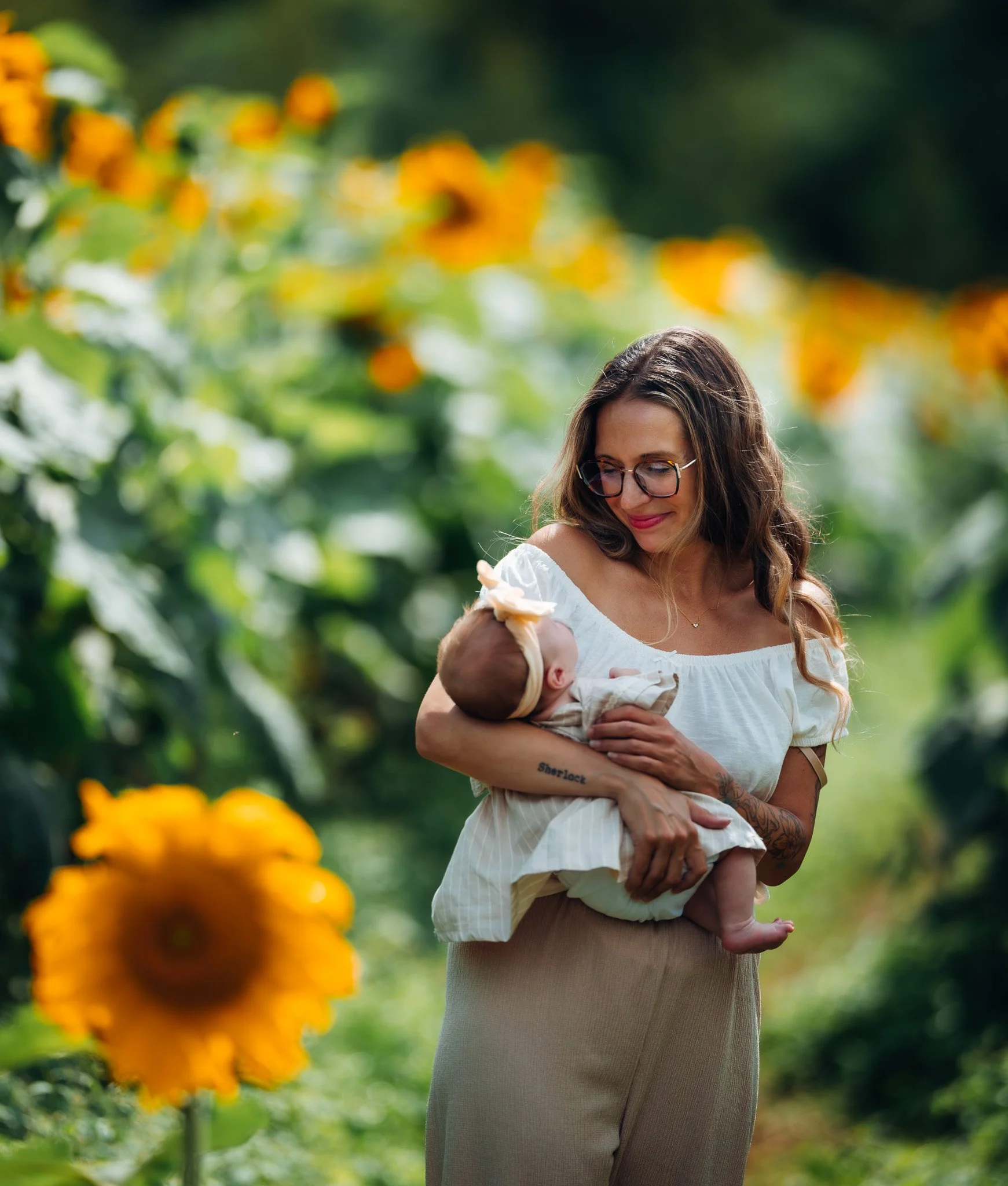 A woman with long brown hair, glasses, and a white off-shoulder top is smiling while holding a baby in her arms outdoors, with a blurred background of green foliage and yellow flowers.