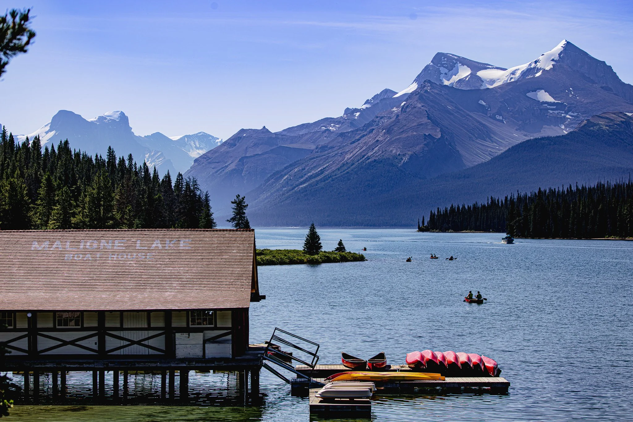 Lake Mona Lake with kayaks and canoes docked, surrounded by pine trees, with mountains in the background and a boathouse in the foreground.