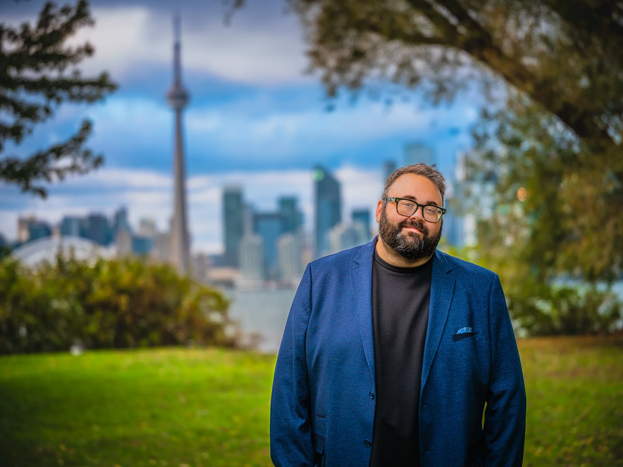 A man with glasses and a beard wearing a blue blazer and black shirt, standing outdoors in a park with the Toronto skyline in the background.