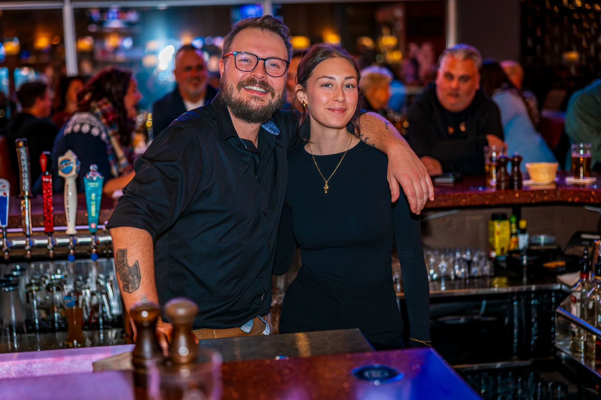A bartender and a woman posing for a photo behind a bar at a lively social gathering. The bartender has glasses and a beard, wearing a black shirt, and the woman has dark hair, a necklace with a cross, and a black outfit. There are other people in th