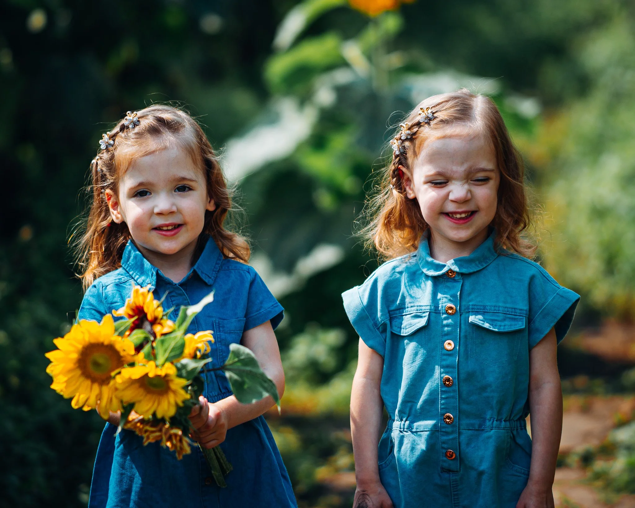 Two young girls with red hair and flower headbands wearing blue dresses, standing outdoors in front of green foliage. One girl is holding a bouquet of sunflowers and other yellow flowers, smiling with her eyes slightly closed, while the other girl is
