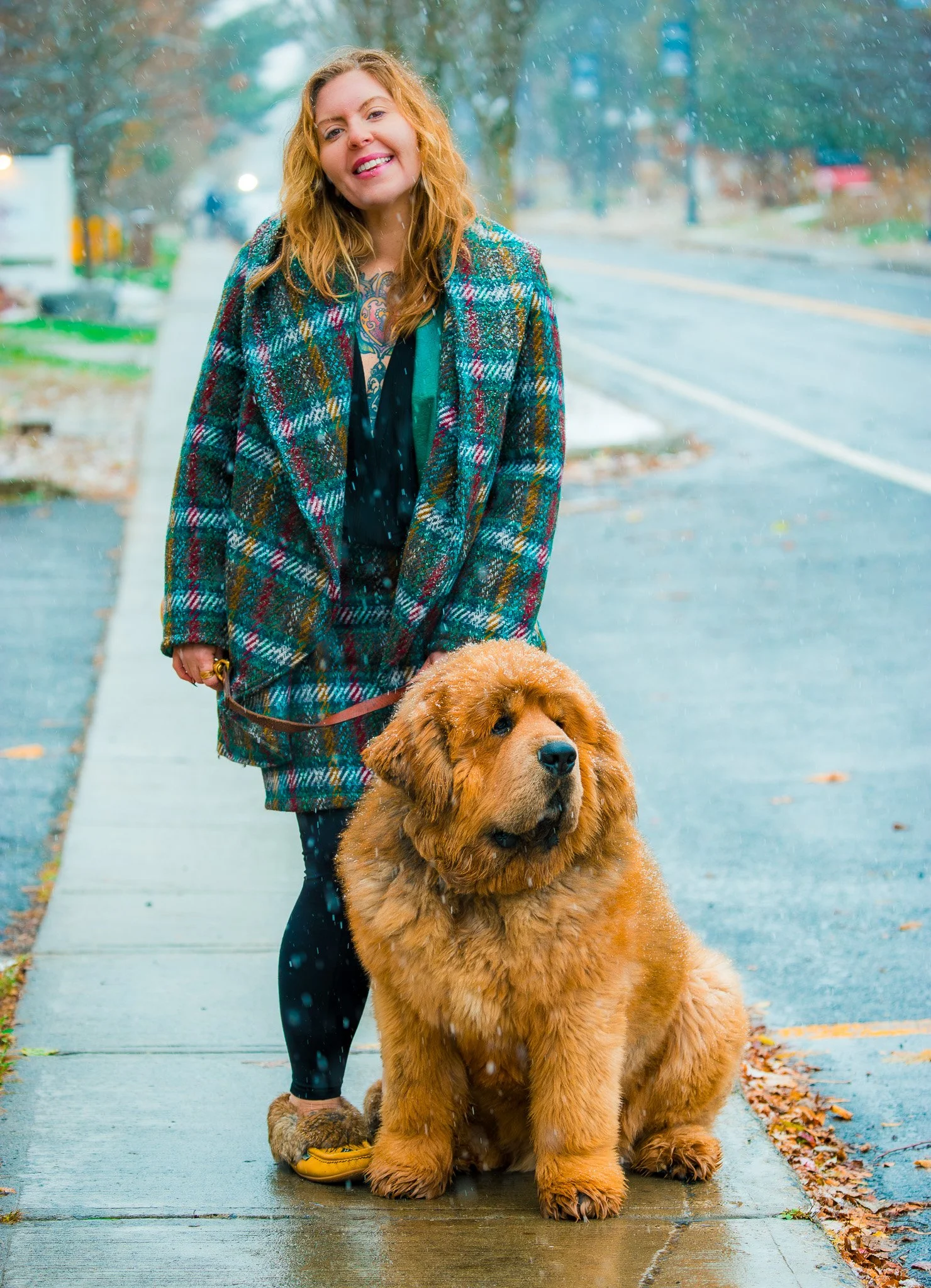 A woman with long red hair smiling while standing on a rainy sidewalk, holding a leash attached to a large, fluffy golden retriever sitting beside her.