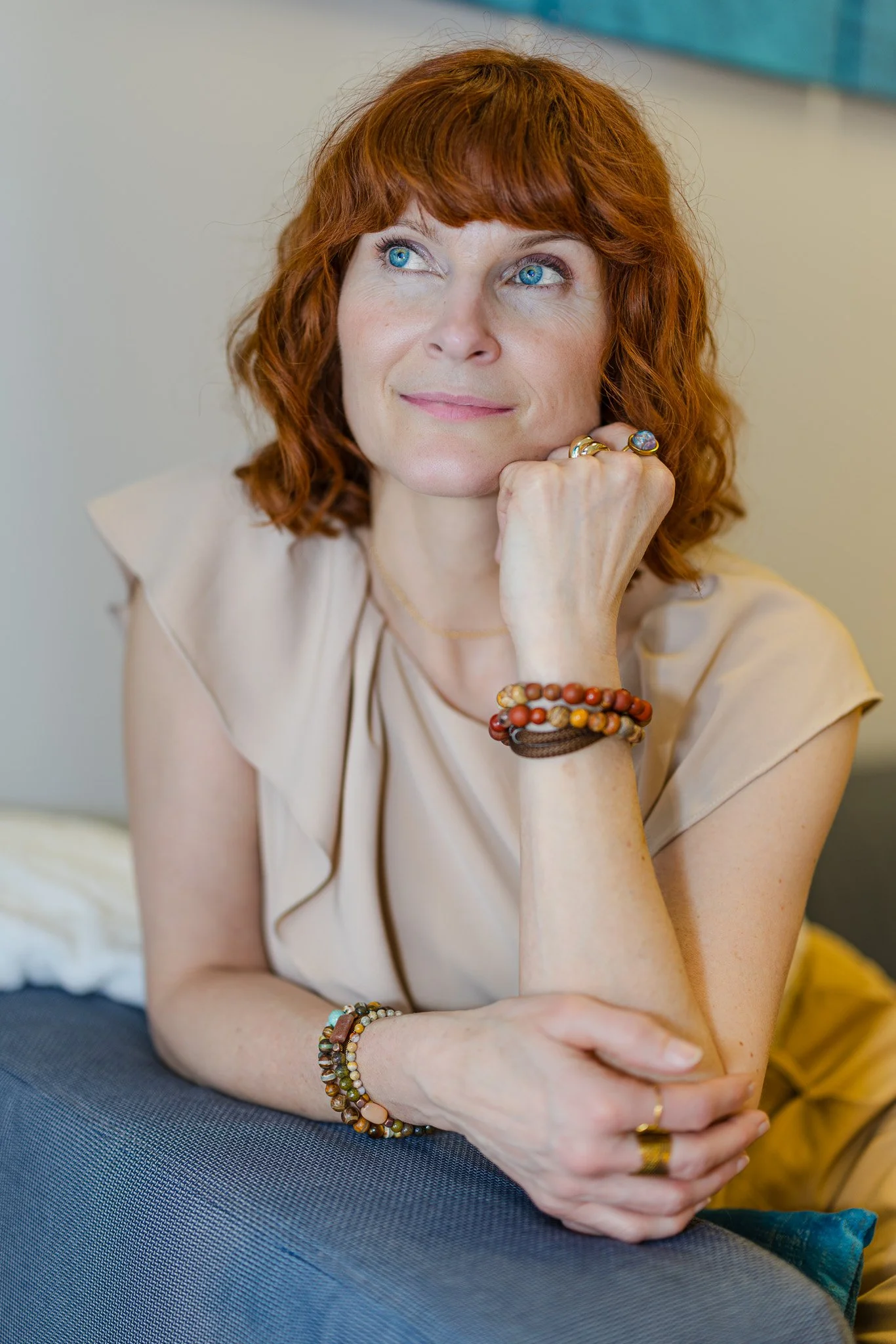 A woman with red hair and blue eyes resting her chin on her hand, wearing rings and bracelets, seated indoors.