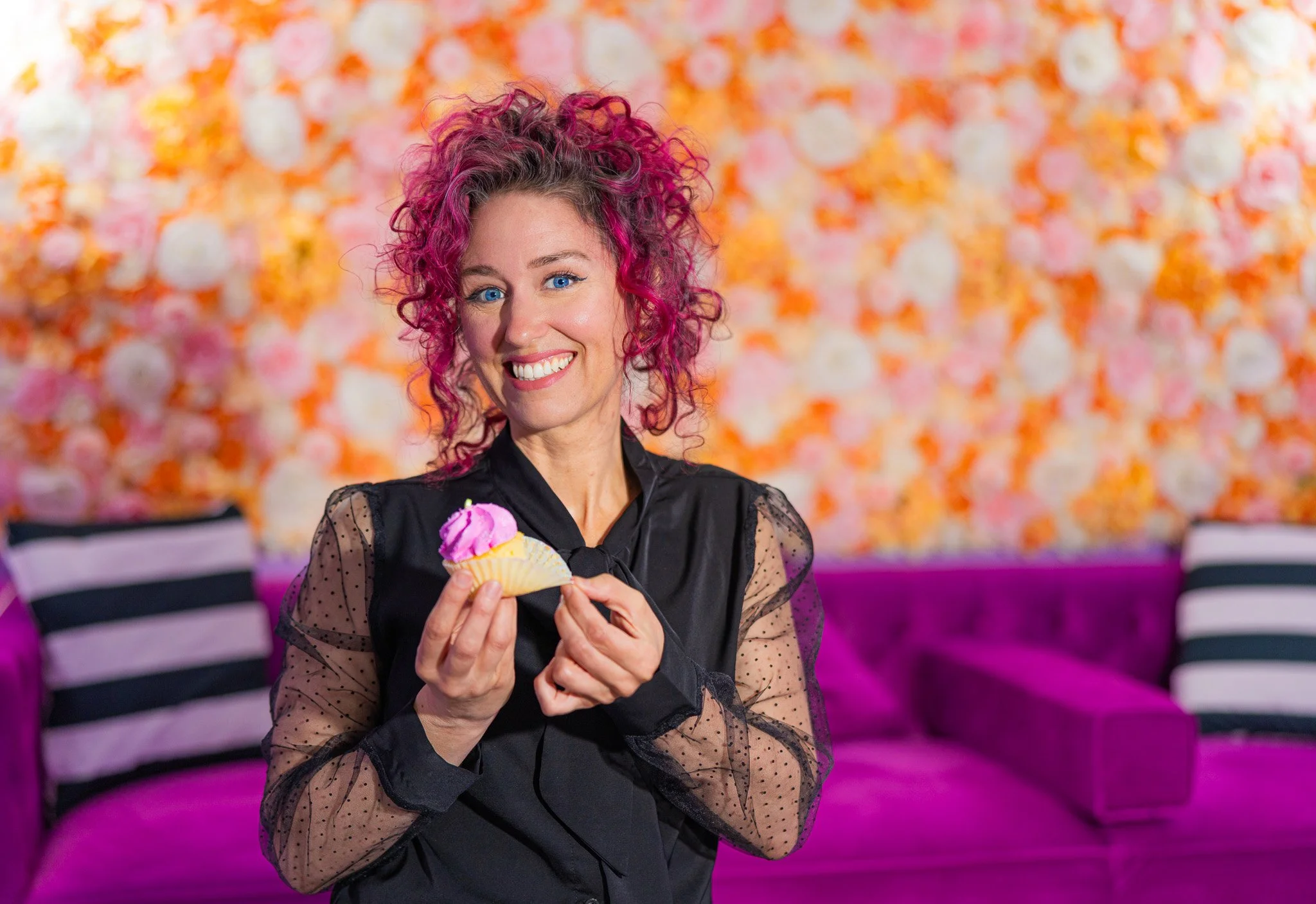 A woman with curly pink hair smiling and holding a cupcake with pink frosting in a colorful, floral-themed room.