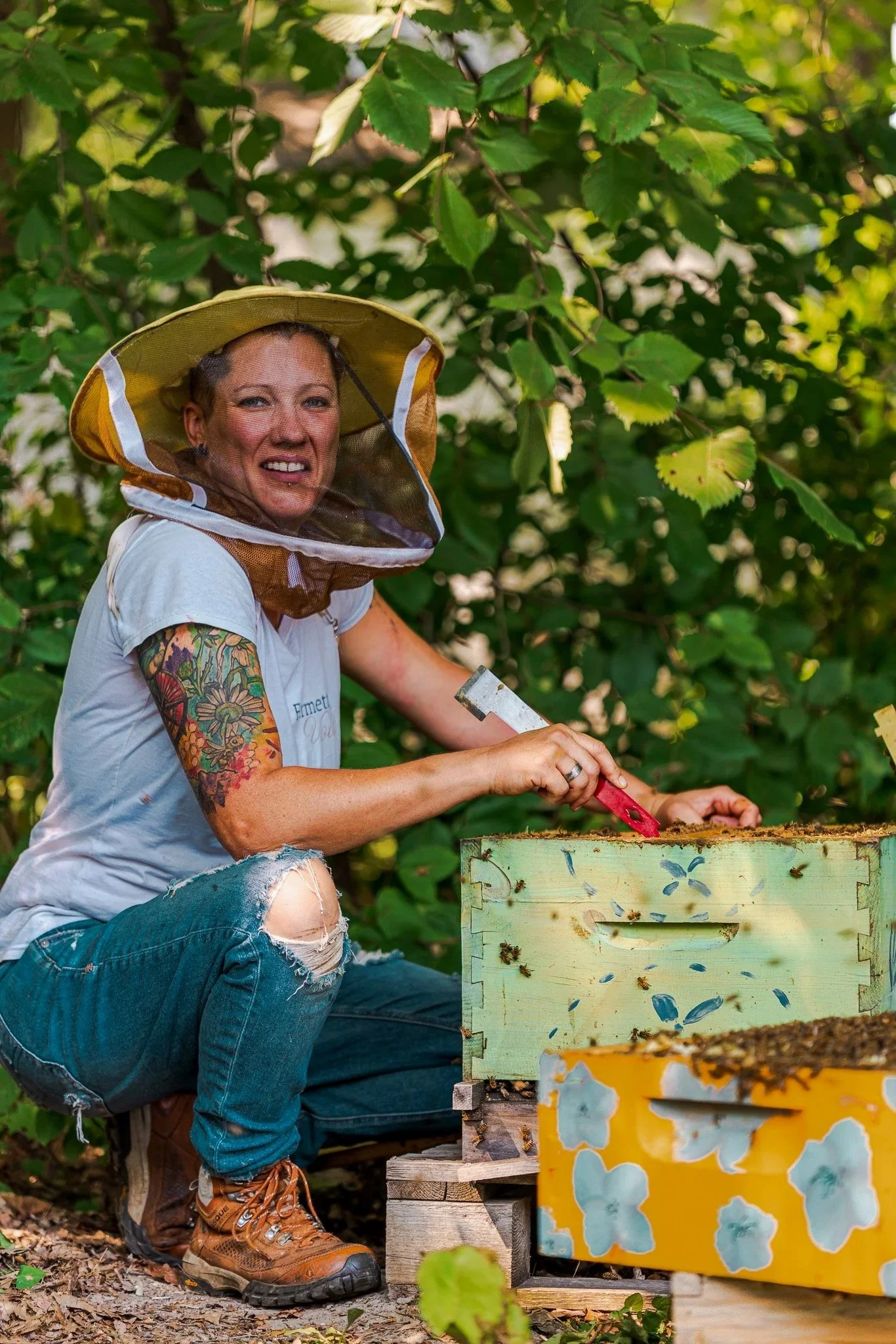 A woman wearing a beekeeping hat and gloves, crouching next to a bee hive, smiling while working with the hive, surrounded by green leaves.