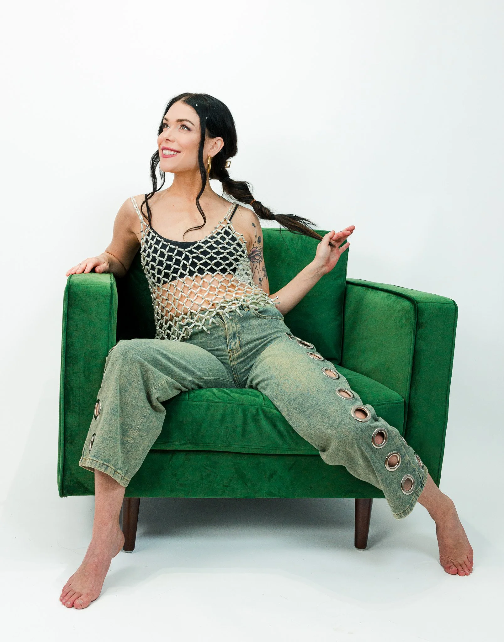 A woman sitting on a green couch, wearing a black and white crochet top and baggy jeans with metal eyelets, smiling and looking to her left.