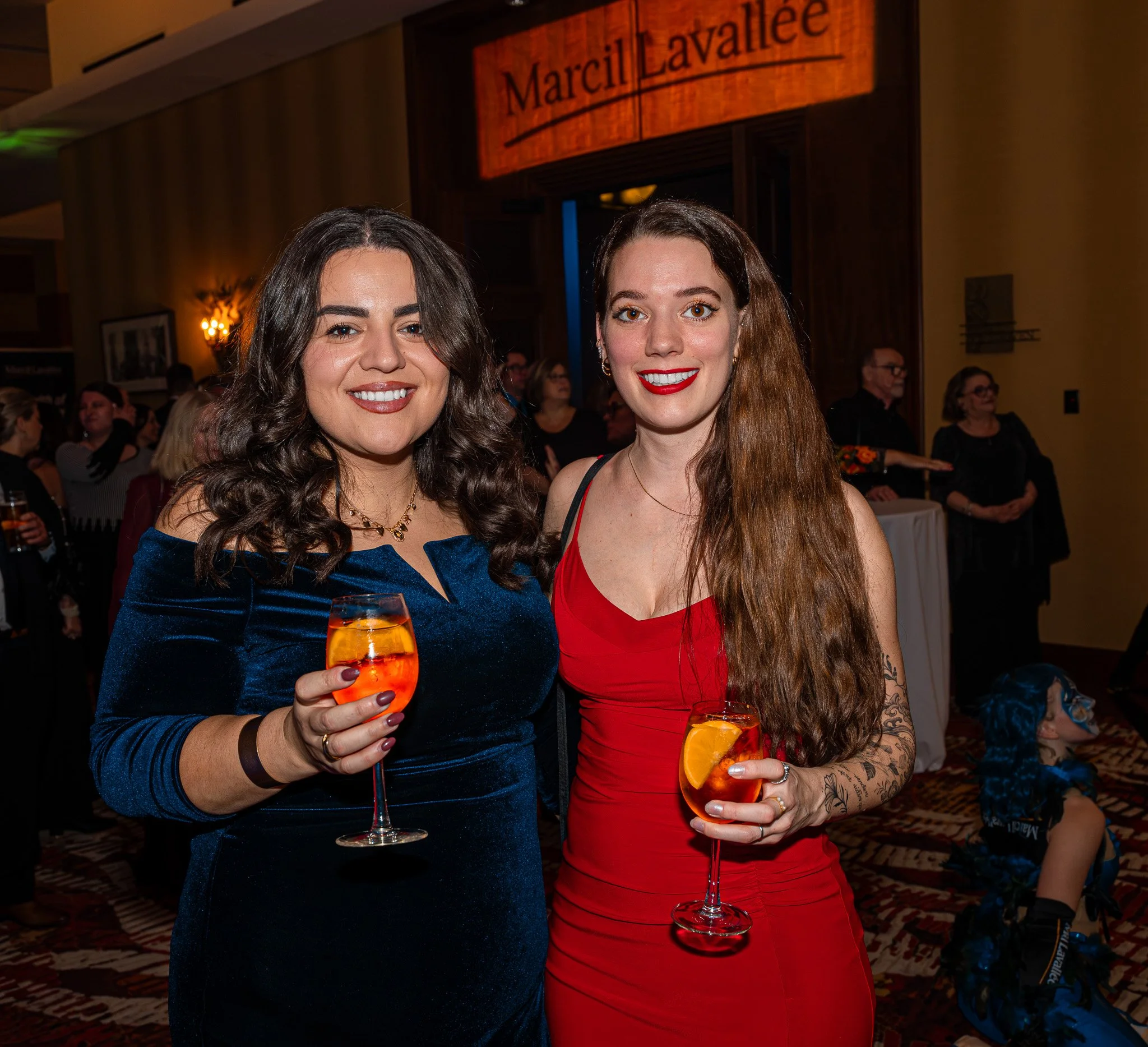 Two women smiling at a party, holding glasses of orange-colored drinks with orange slices, standing in a decorated indoor event space.