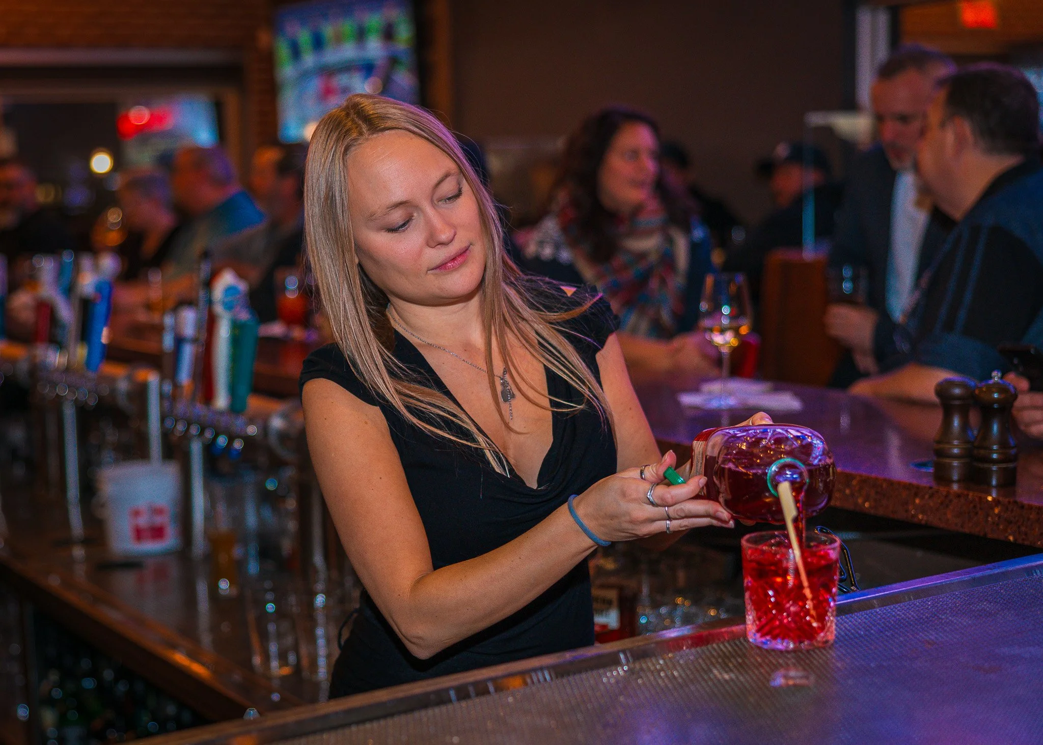 A woman standing at a bar, pouring a red drink from a large glass pitcher into a glass. She is wearing a black top and is in a lively, crowded bar with other patrons in the background.