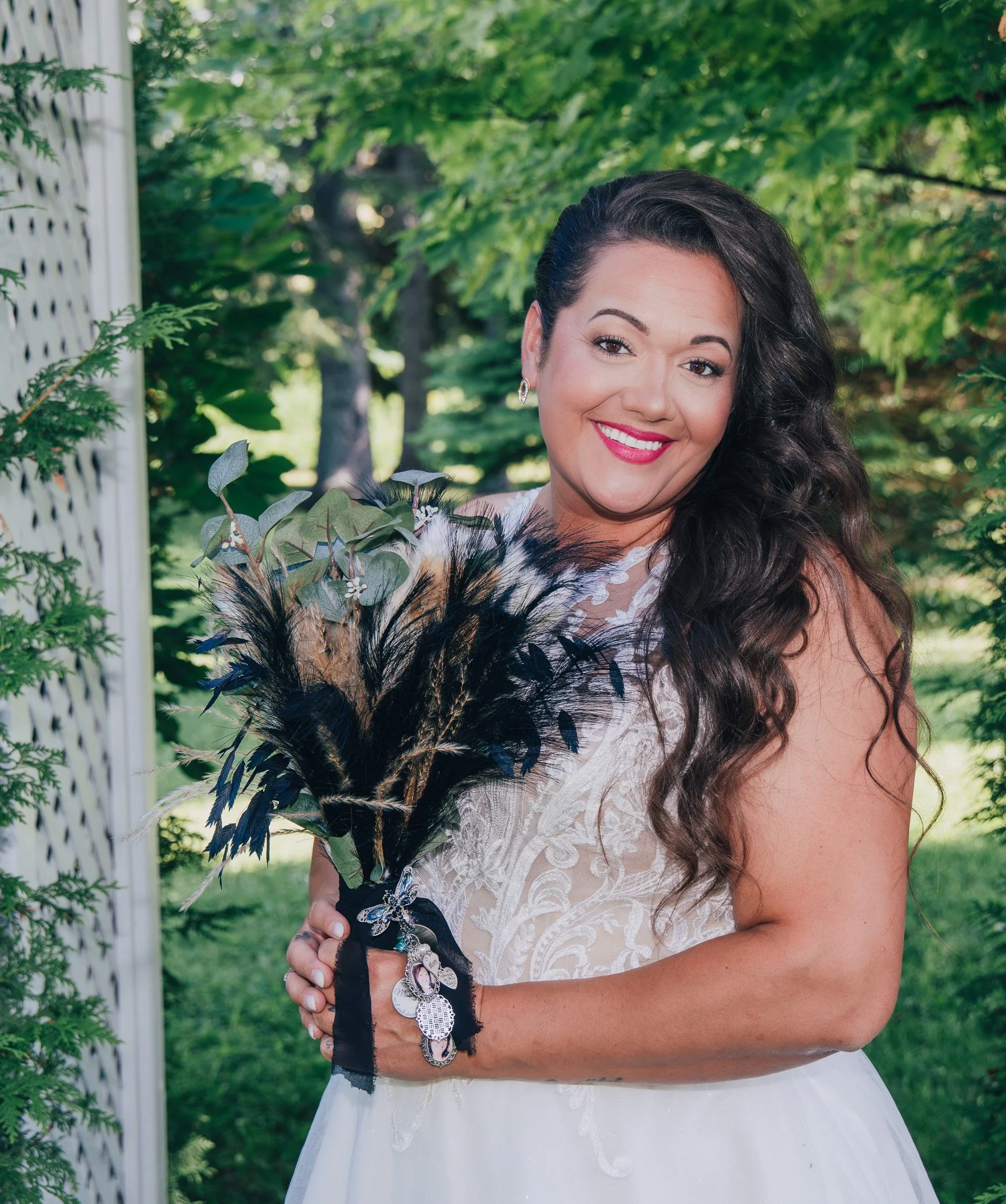A woman with long curly hair in a white dress holding a bouquet of feathers and greenery, standing outdoors with green trees in the background.