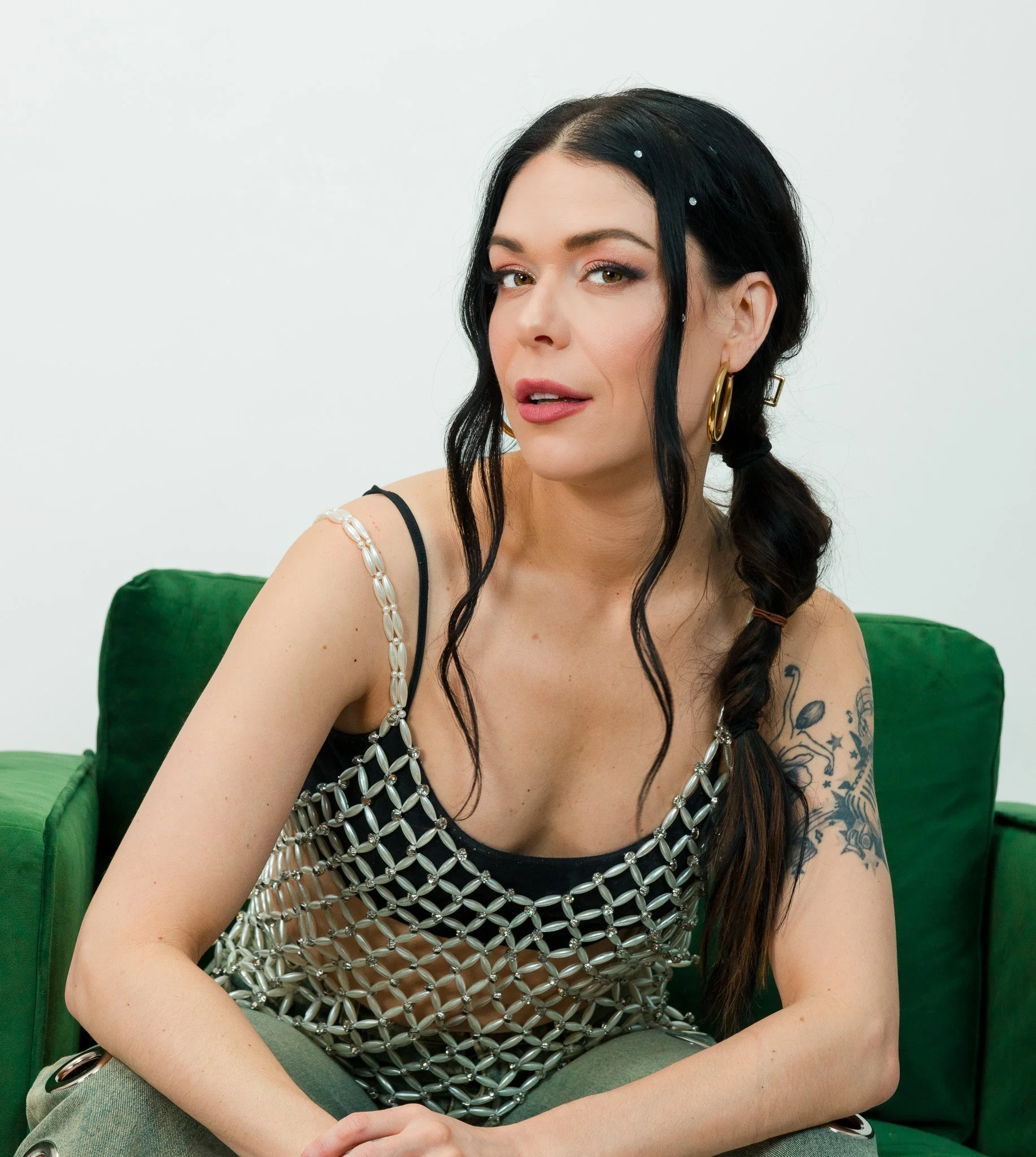 A woman with fair skin, black hair, and tattoos on her right arm is sitting on a green chair, looking at the camera with a slight smile. She is wearing large gold earrings, a black tank top with a silver chainmail overlay, and has colorful makeup.