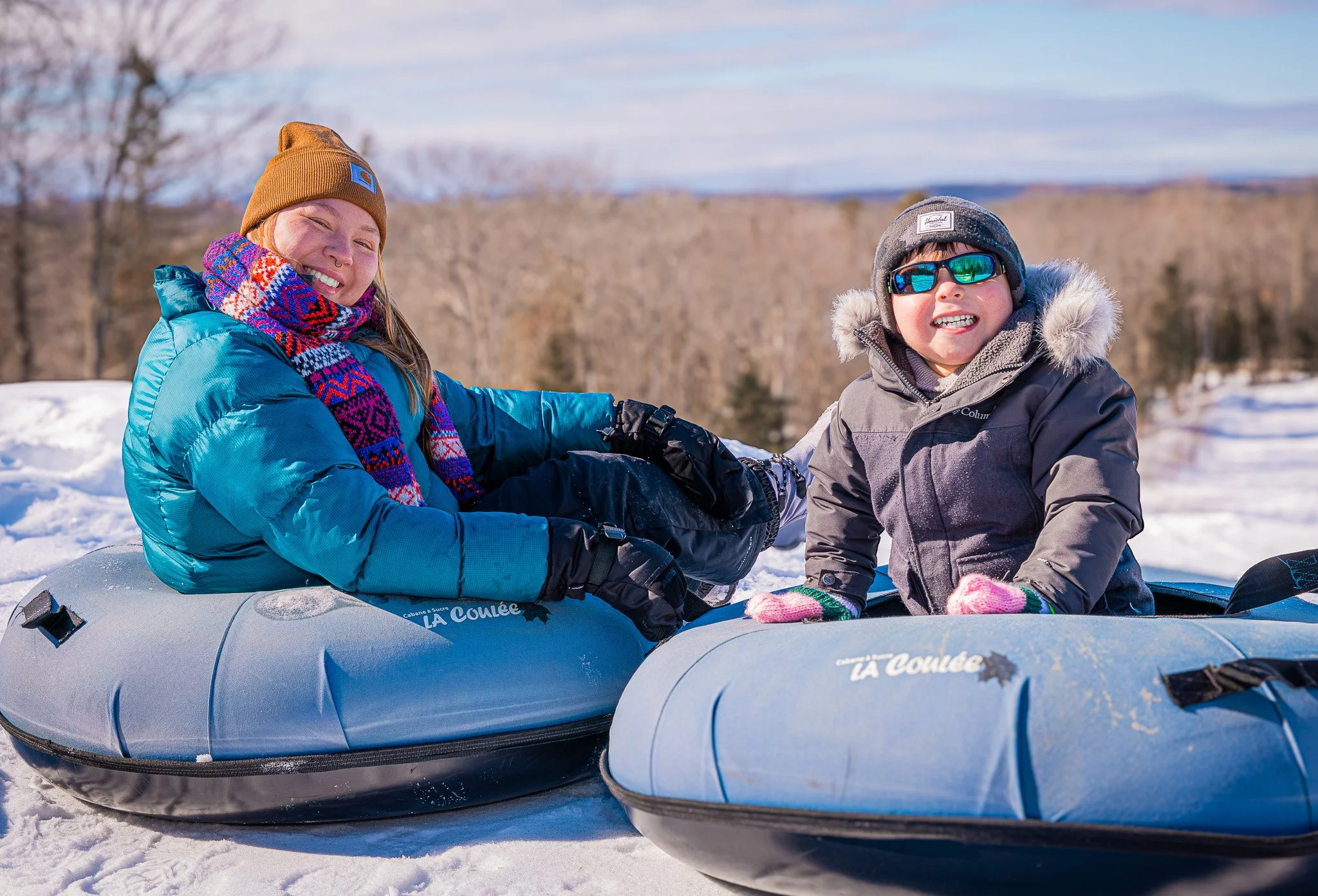 A woman and a young boy dressed in winter gear sitting on inflatable snow tubes in a snowy outdoor landscape.