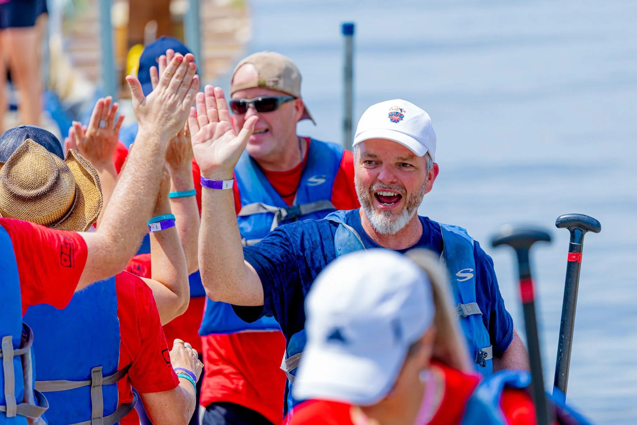 Group of people at a marina high-fiving and smiling, dressed in casual outdoor clothing and life jackets, with boats and water in the background.