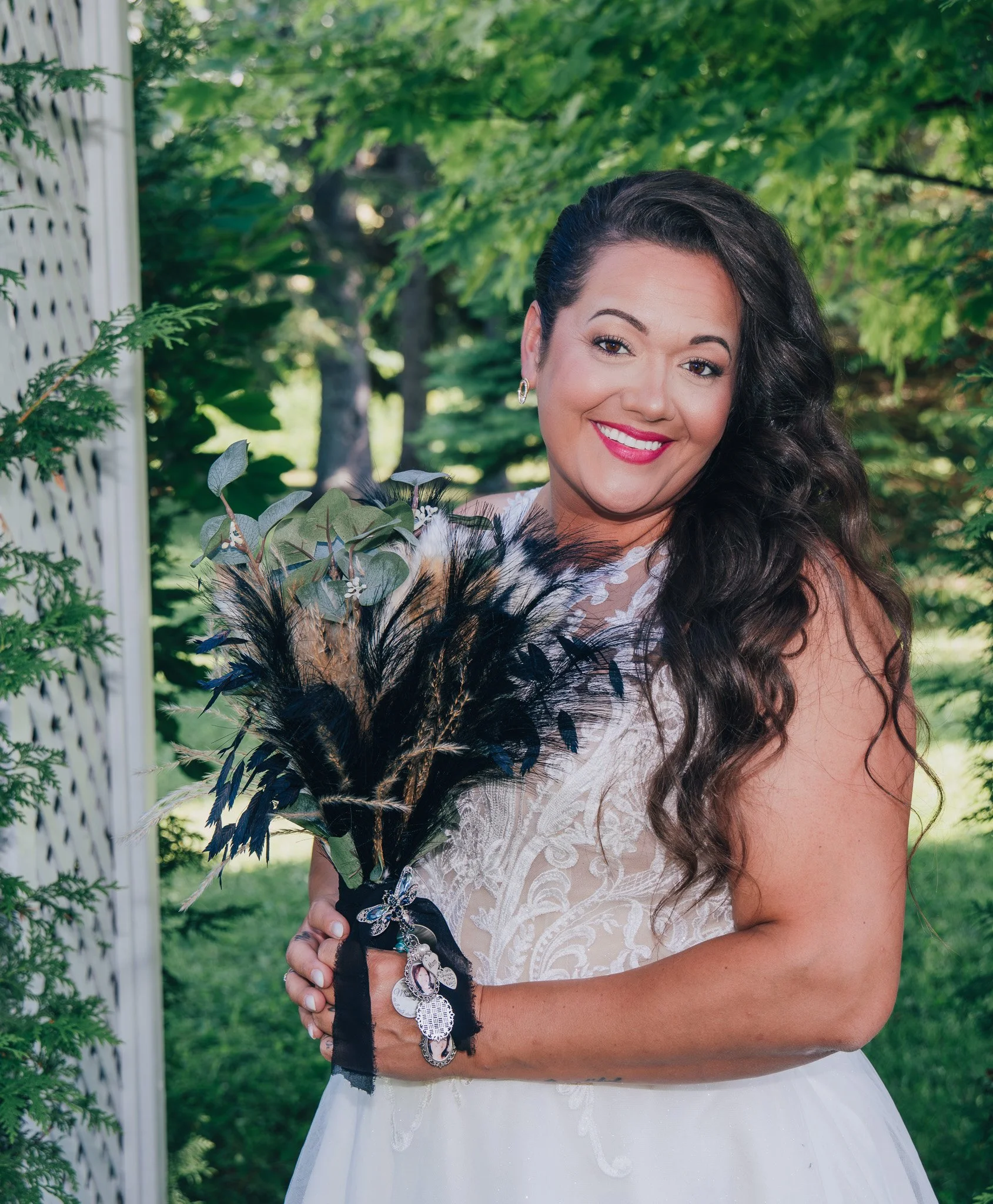 A woman with long, dark, curly hair smiling outdoors in a white dress, holding a bouquet of black and brown feathers, greenery, and charms.