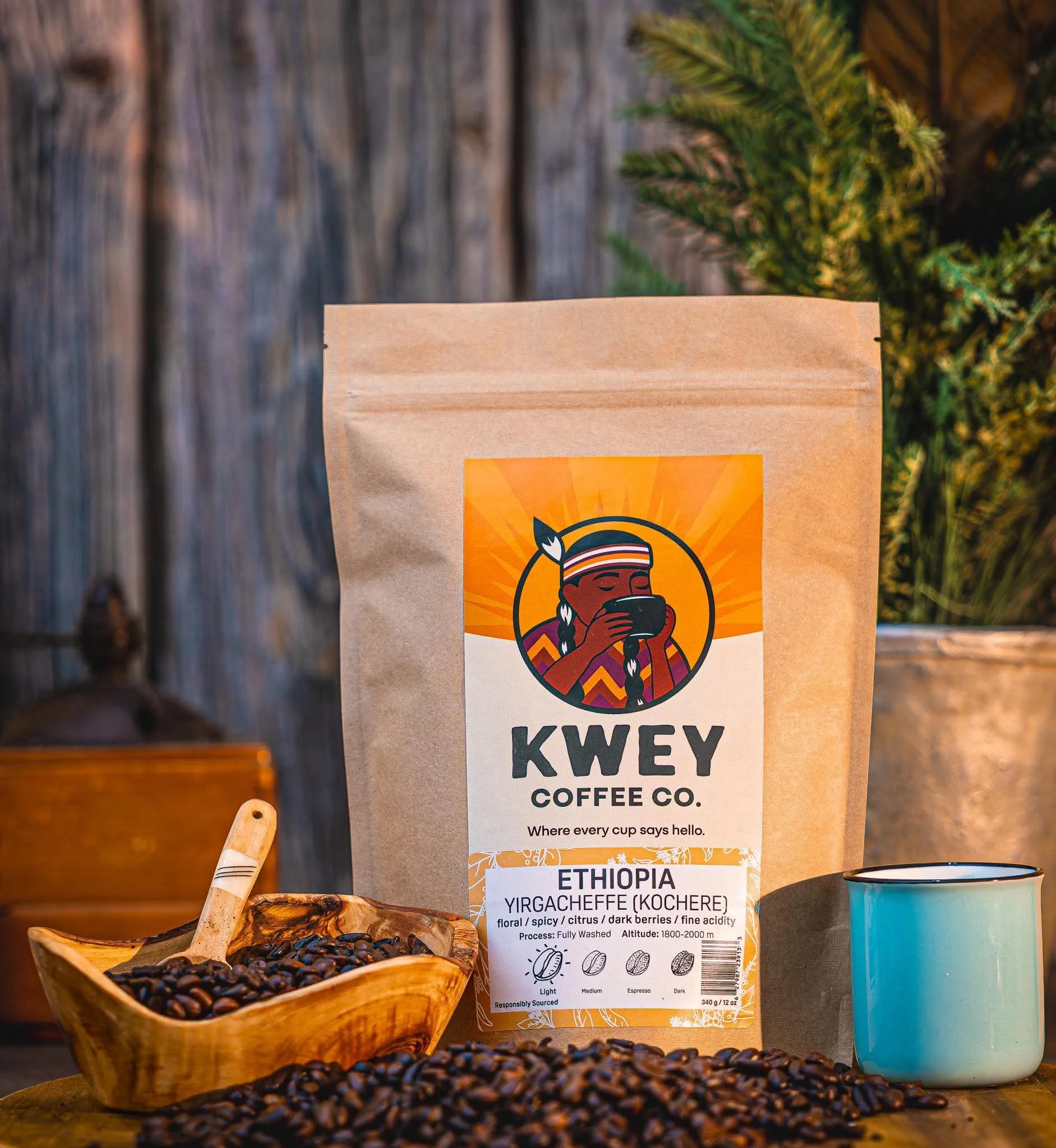 A bag of Kwey Coffee Co. Ethiopia Yirgacheffe coffee beans on a wooden table, with coffee beans in a wooden scoop and scattered on the table, alongside a mug and a greenery background.