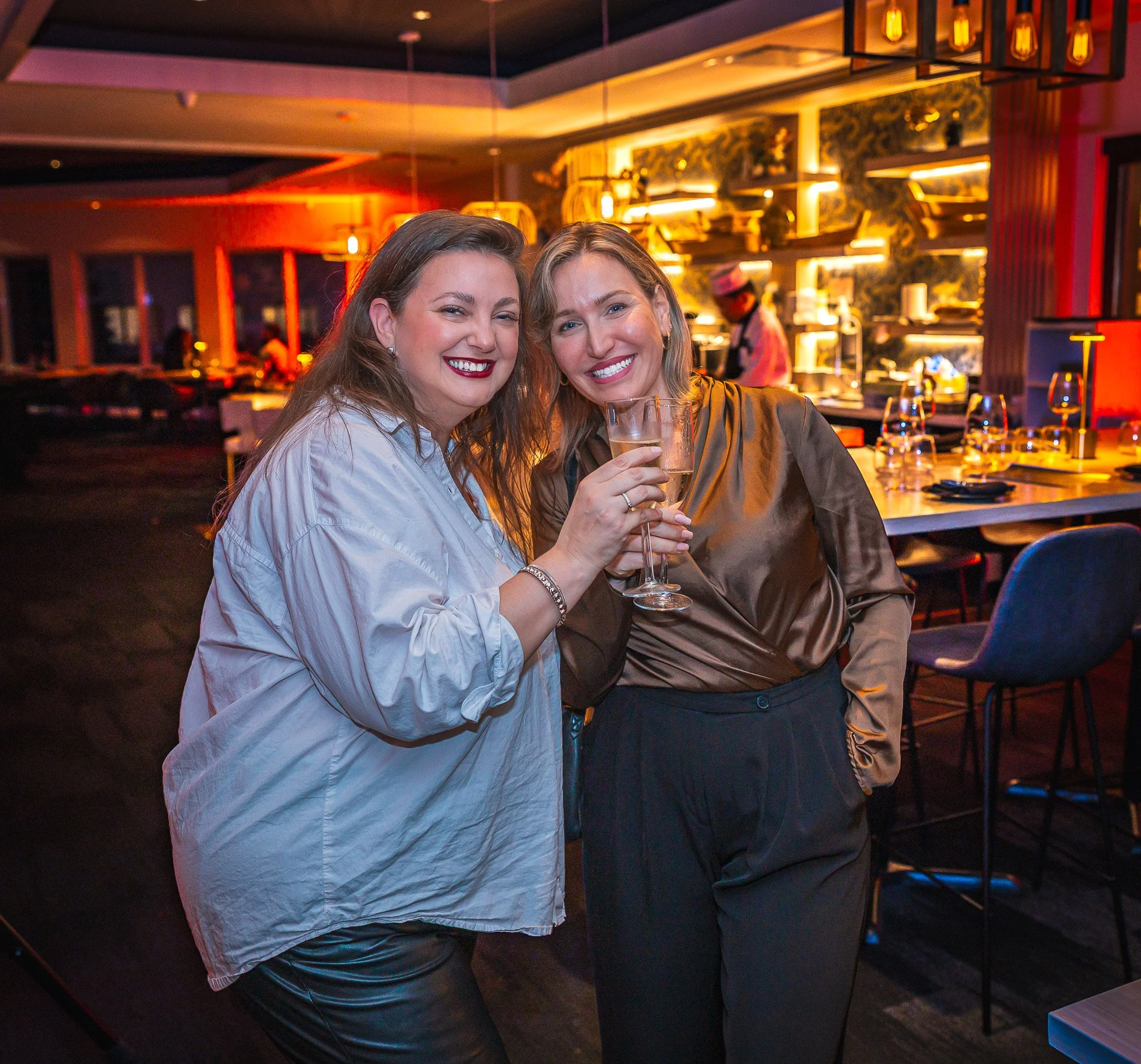 Two women smiling and holding glasses of champagne in a restaurant with warm lighting and a lively atmosphere.