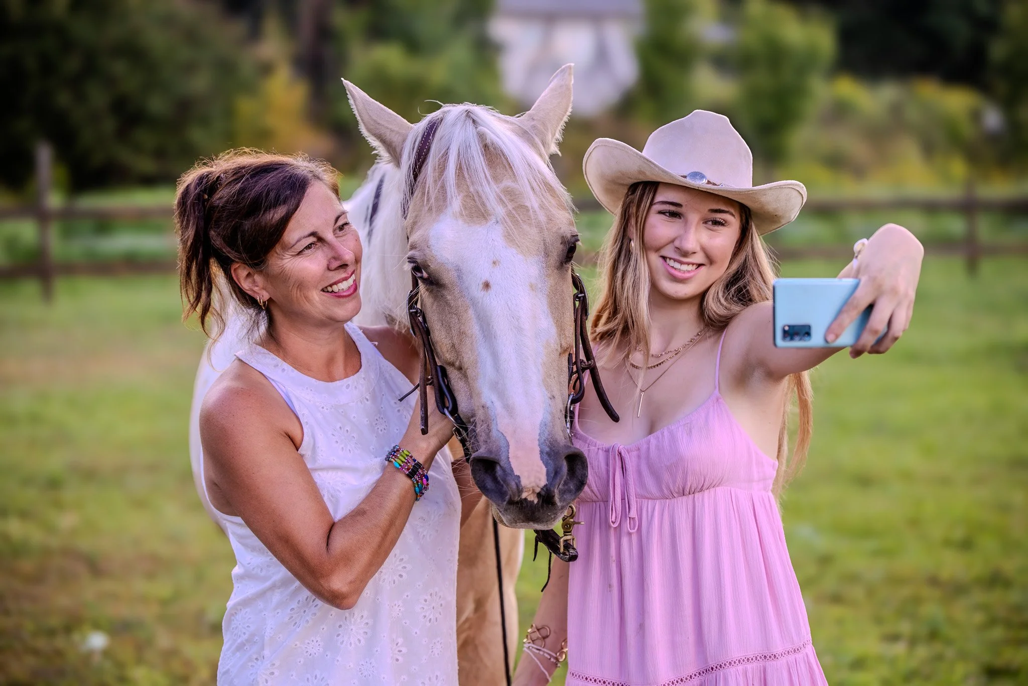 Two women, one middle-aged and one young, taking a selfie with a white horse in a green outdoor field. The older woman is smiling and holding the horse's bridle, while the young woman is wearing a cowboy hat and holding the phone.