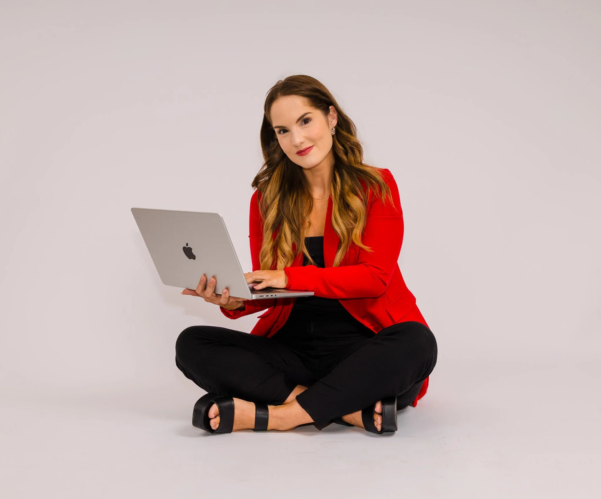 Woman sitting cross-legged on the floor holding an open MacBook laptop, wearing a red blazer, black top, black pants, and black sandals, against a plain light background.