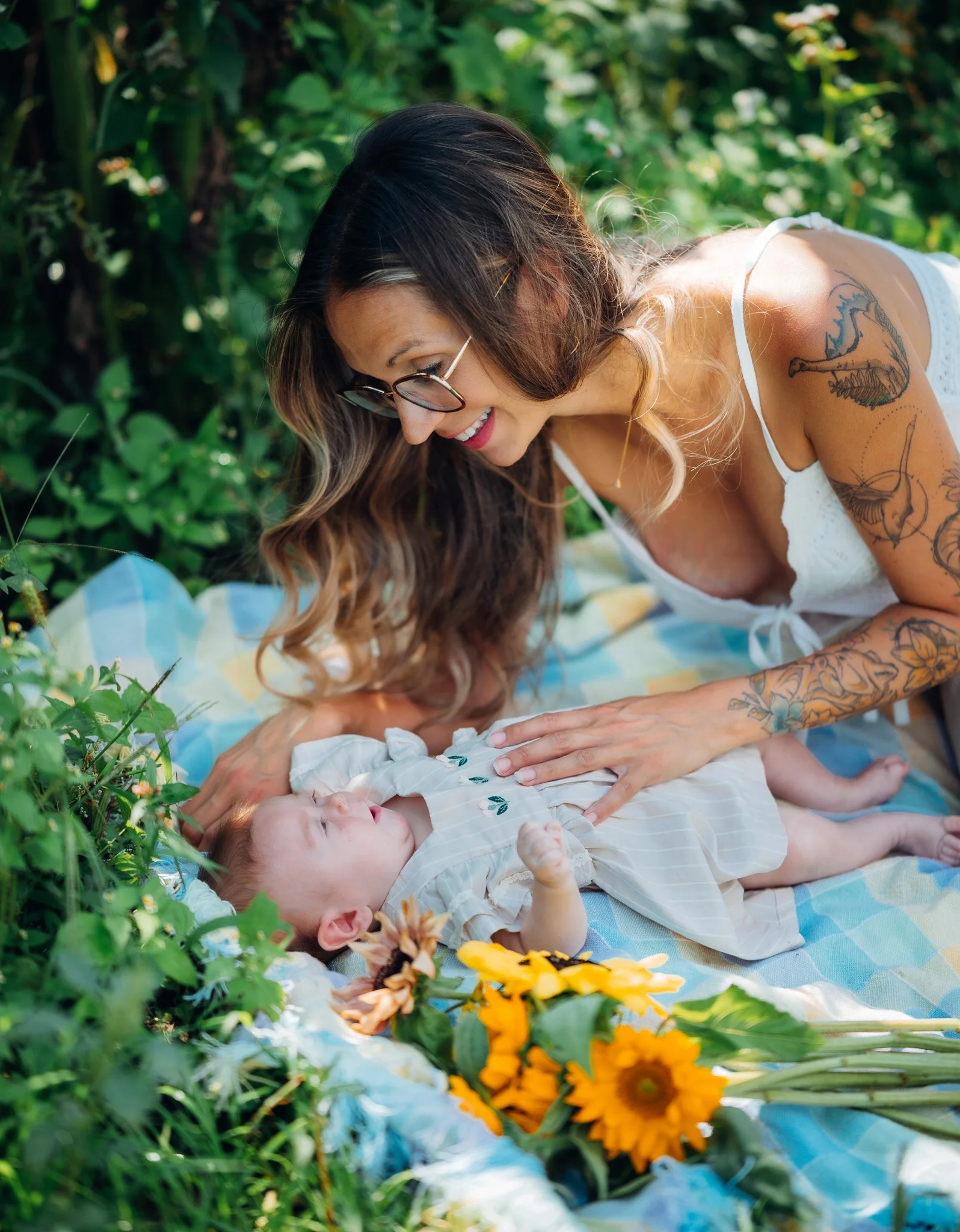 A woman with glasses, long hair, and tattoos, smiling and looking down at a baby lying on a blanket outdoors in a garden surrounded by green plants and yellow flowers.