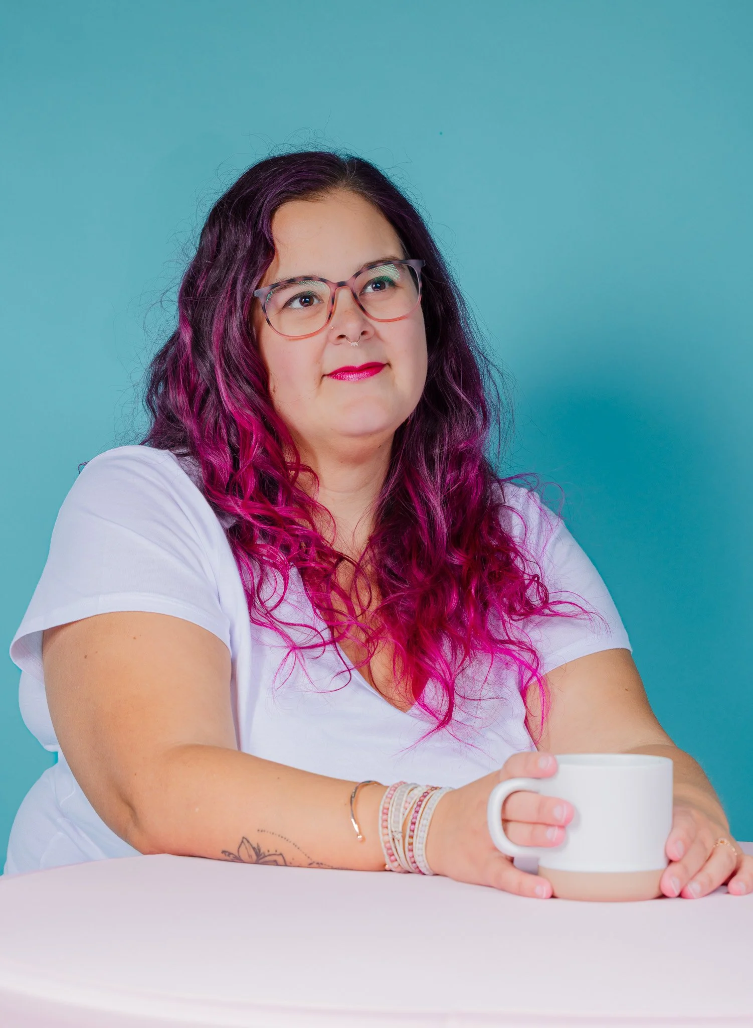 A woman with long, curly purple-pink hair and glasses sitting at a table holding a white mug, with a turquoise background.
