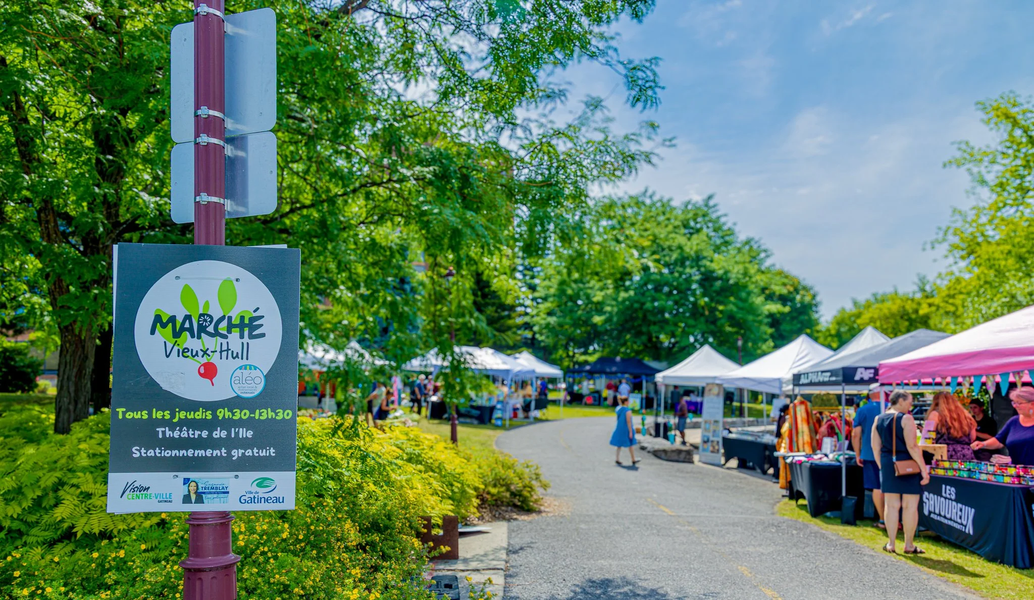 Outdoor marketplace with white tents and vendors, people browsing, green trees, and a sign for the Old Hull Market in Gatineau.