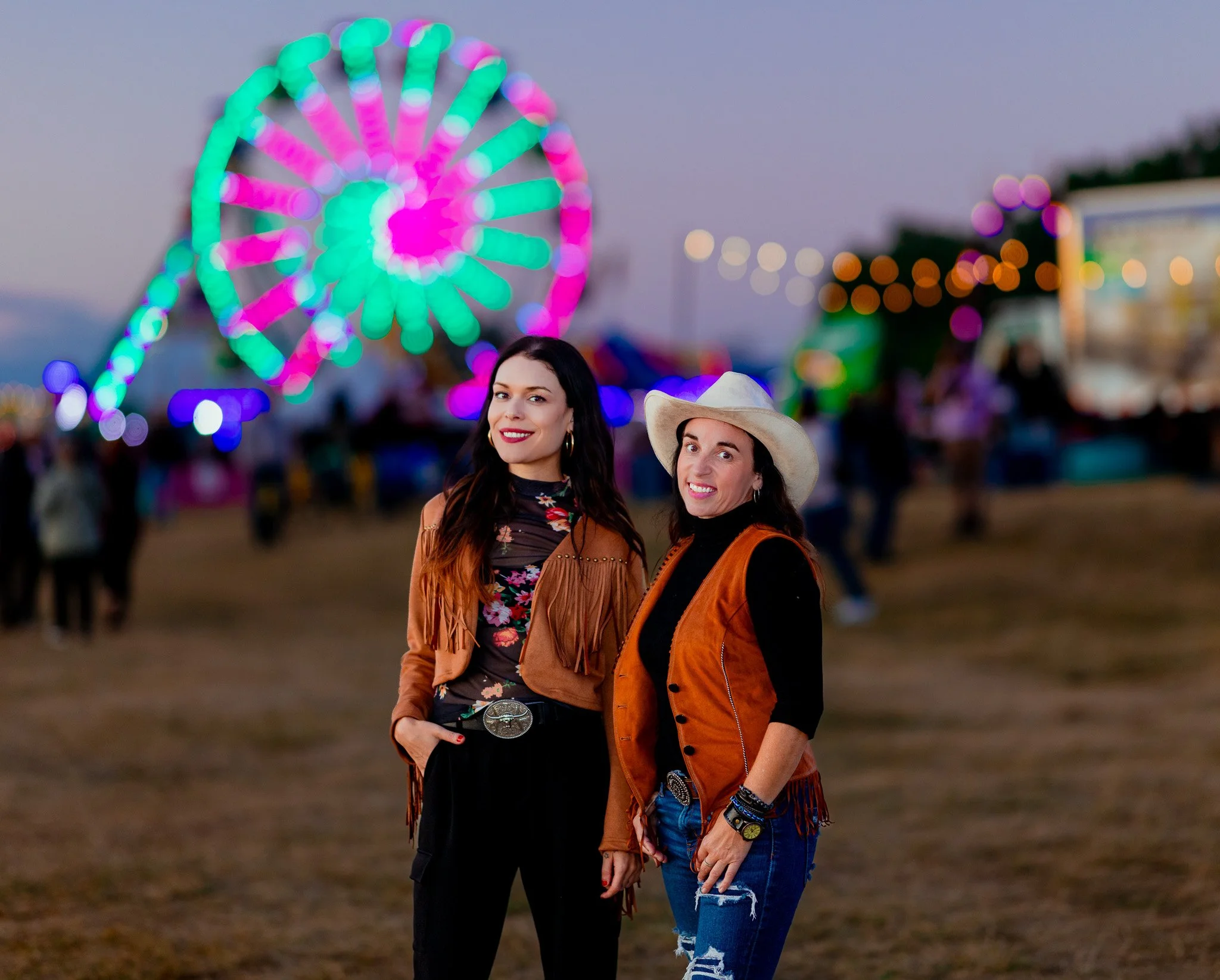 Two women standing at a fairground during twilight, with a large colorful illuminated Ferris wheel in the background. They are smiling and wearing casual festival attire, including cowboy hats and fringed vests.