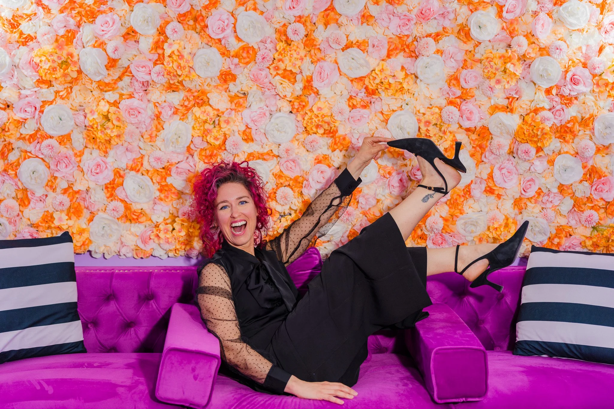 A woman with pink curly hair laughing and posing on a pink velvet sofa with a colorful flower wall background, wearing a black dress with sheer polka-dot sleeves and black high heels.