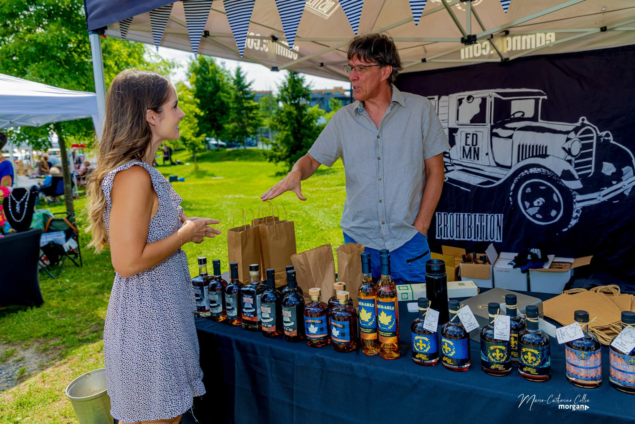 A man and woman are talking at a market stall with bottles of maple syrup. The woman is wearing a sleeveless dress and the man is in a short-sleeve shirt. The stall is under a canopy with a banner displaying a vintage truck that reads 'ED MN' and 'PR