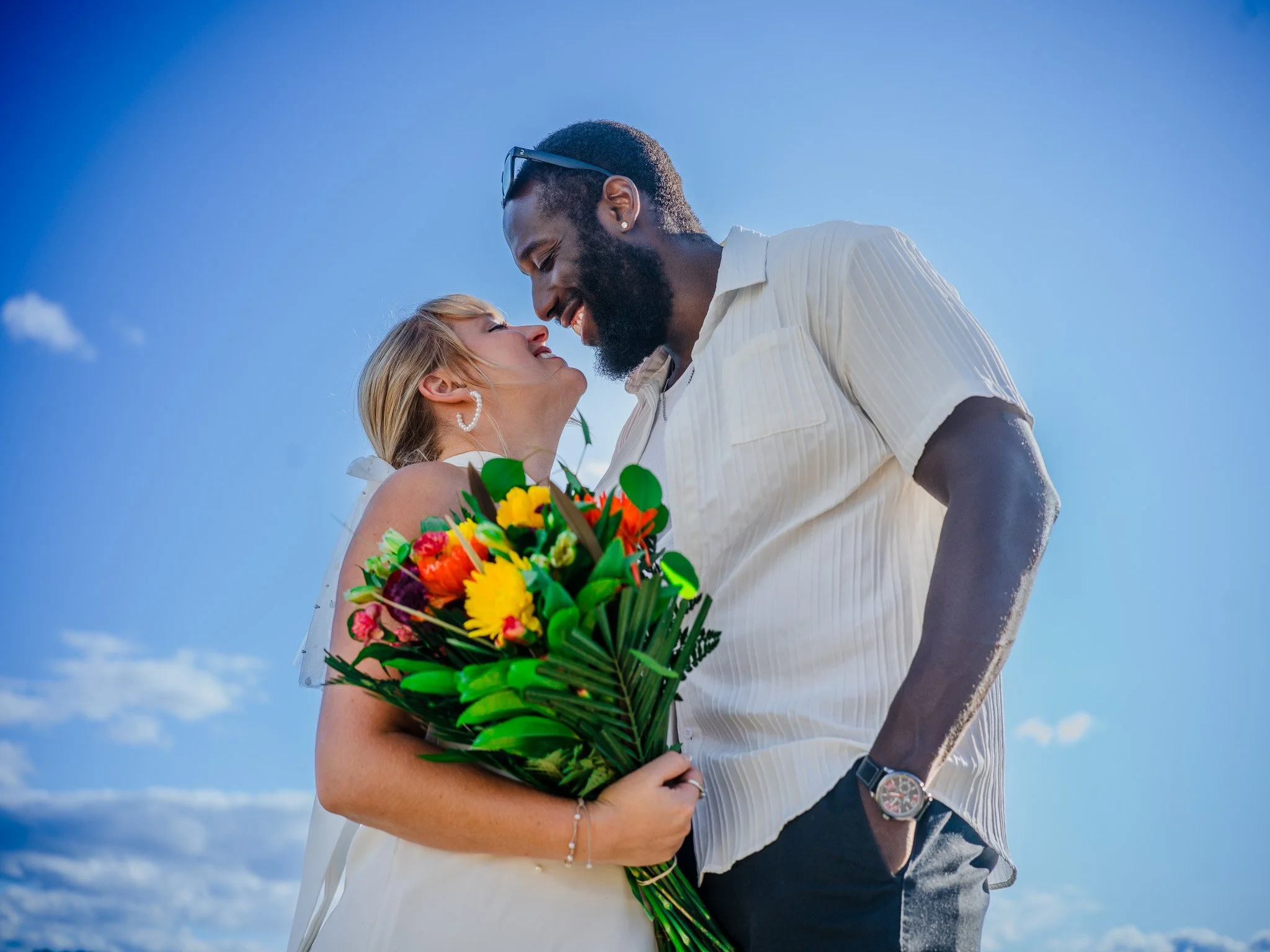 A couple standing close together outdoors on a bright day, the woman holding a colorful bouquet of flowers and the man wearing a watch and casual shirt, as they smile and lean their foreheads together.