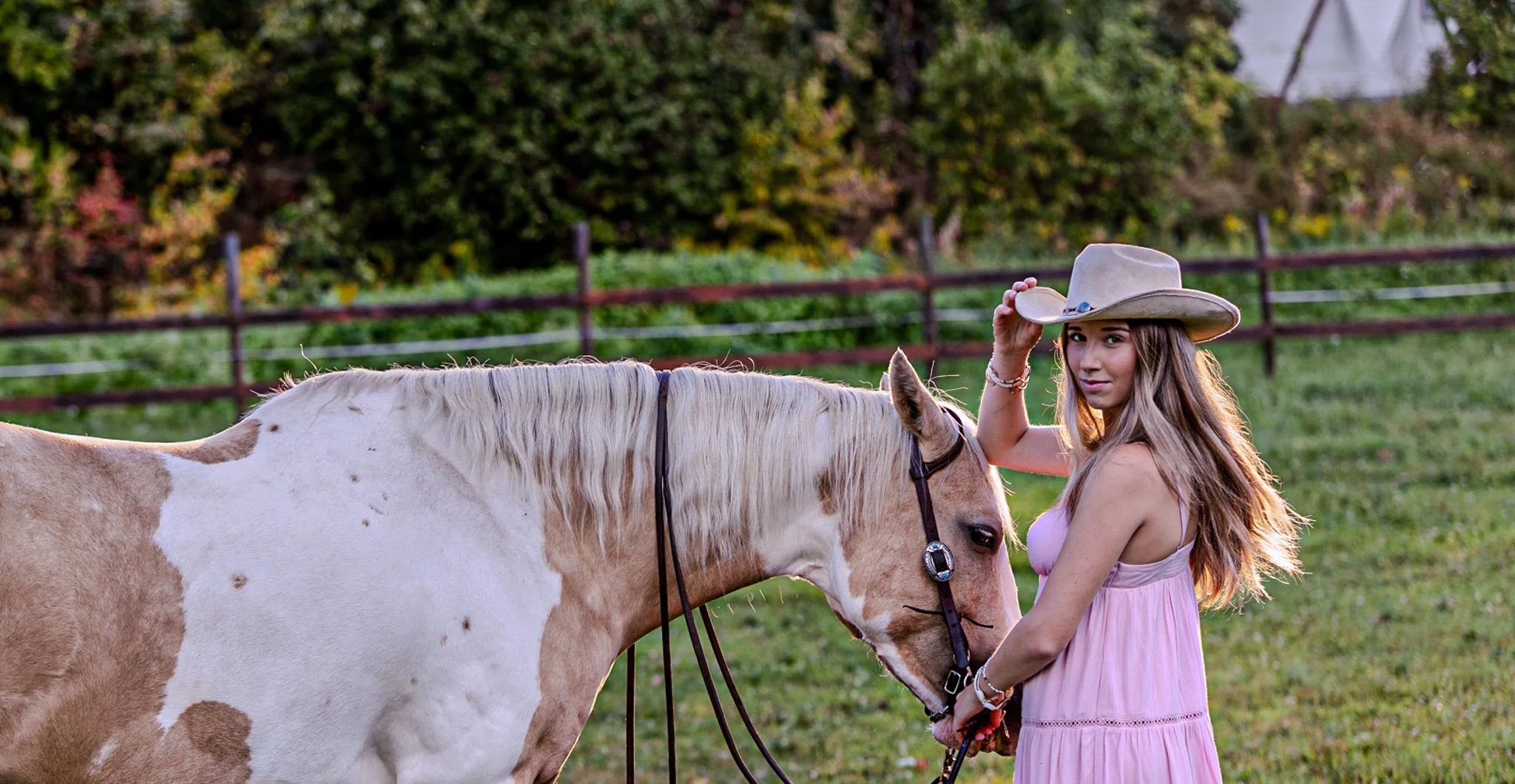 Young woman in pink dress and cowboy hat petting light brown and white horse in green field with trees and a wooden fence in background