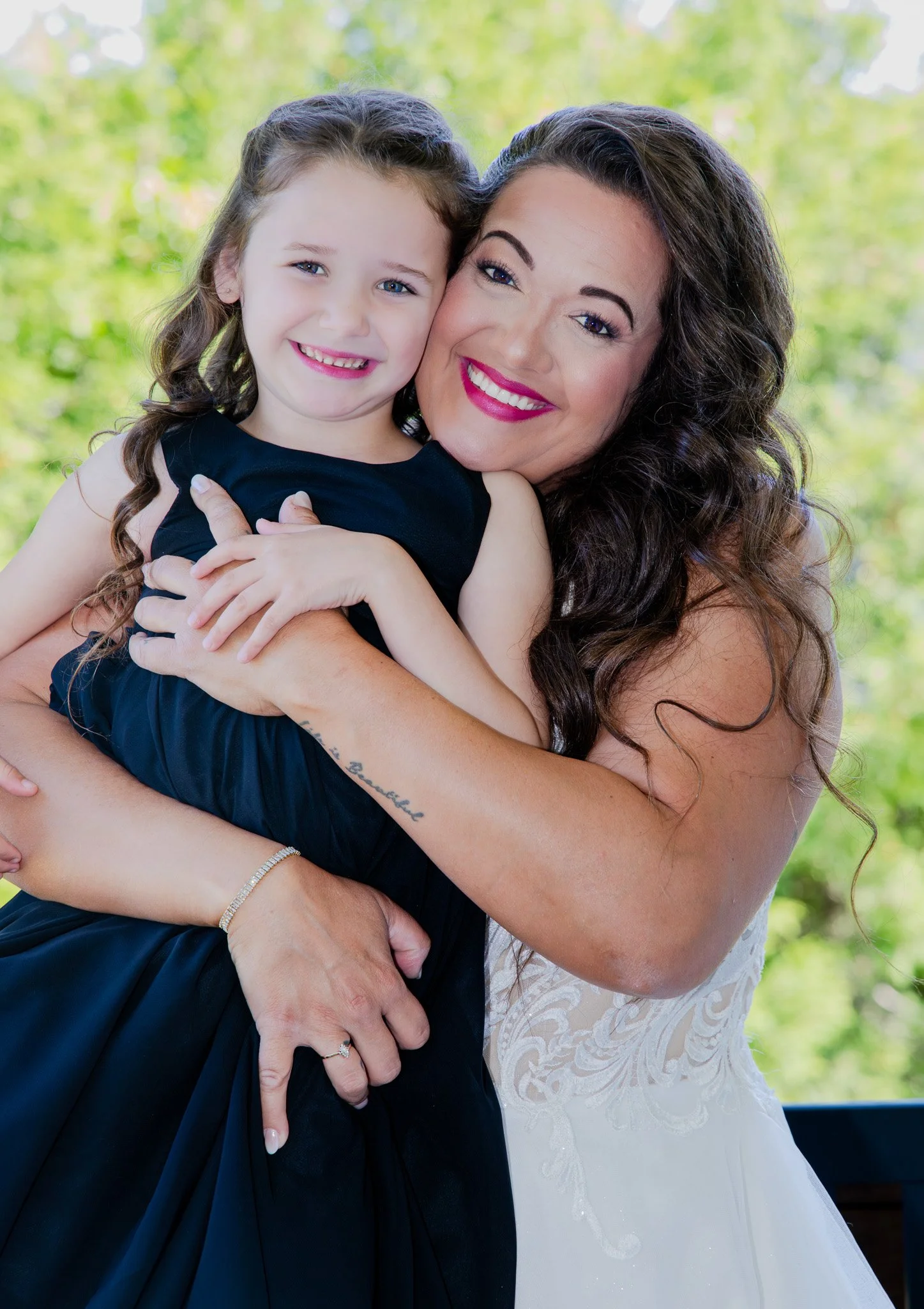 A woman in a white dress hugging a young girl in a black dress outdoors with green trees in the background.