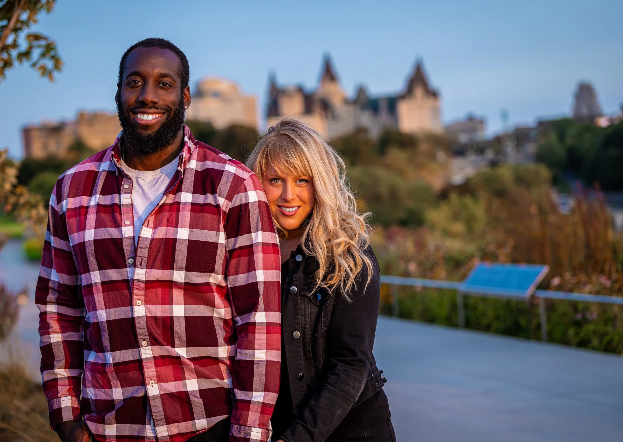 A smiling man in a red plaid shirt and a woman with blonde curly hair in a black jacket pose outdoors with a castle-like building in the background during sunset.