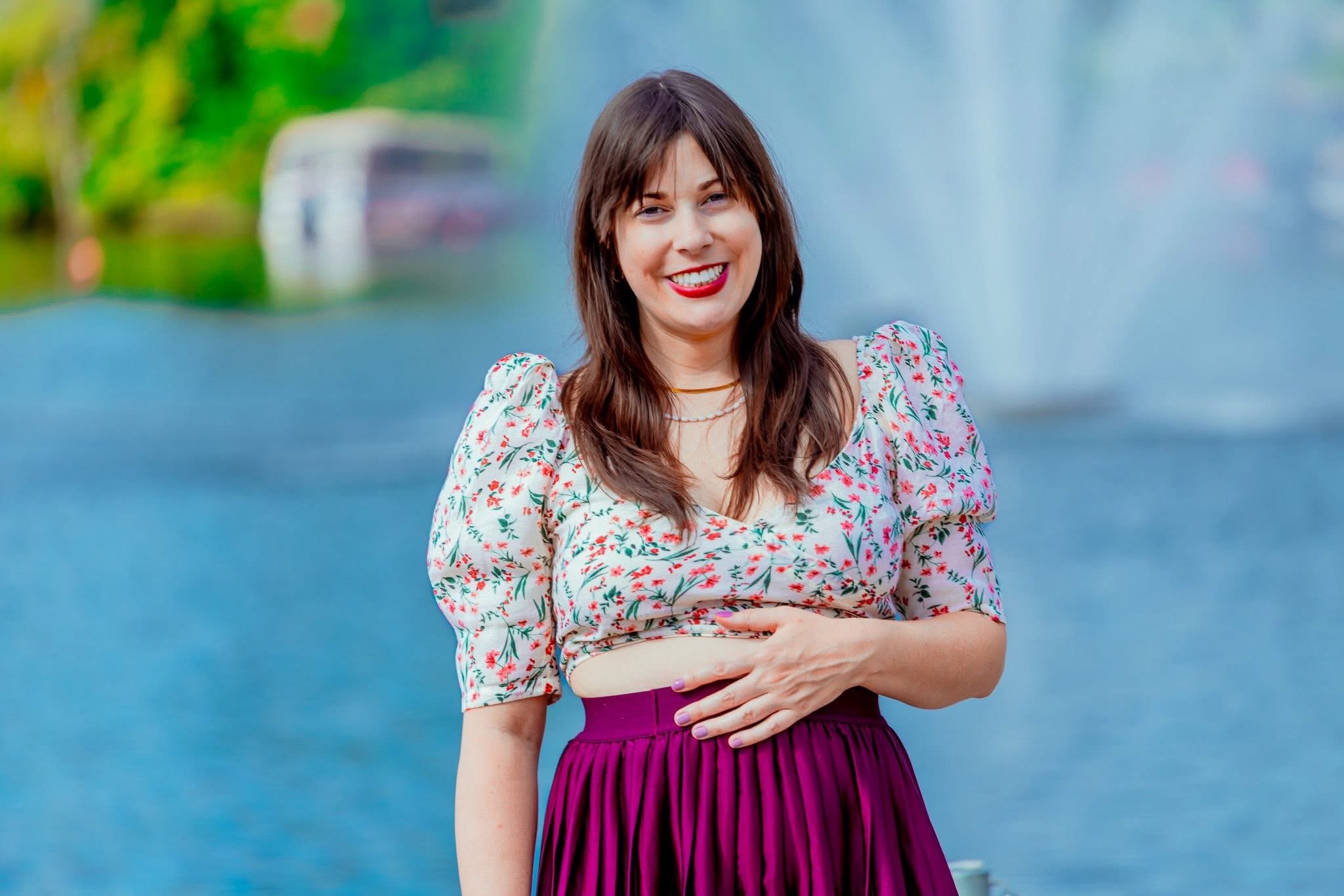 A woman with long brown hair wearing a floral top and a purple skirt, smiling in front of a fountain with water jets and greenery.