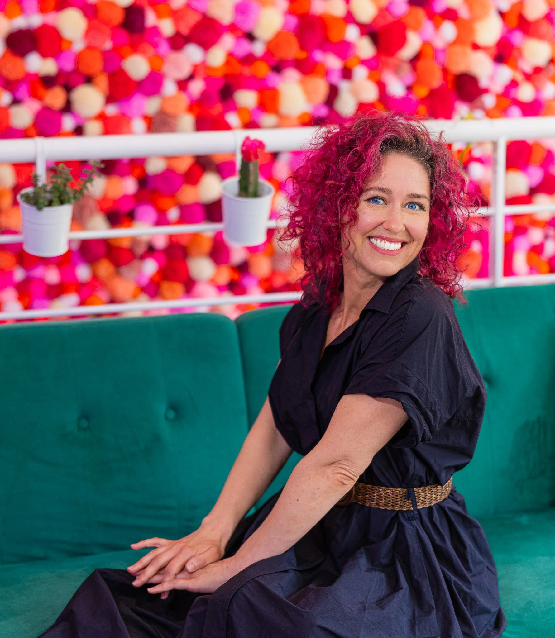 A woman with curly pink hair sitting on a teal sofa, smiling, with pink, orange, and cream pom-poms in the background and small potted plants hanging from a white railing.