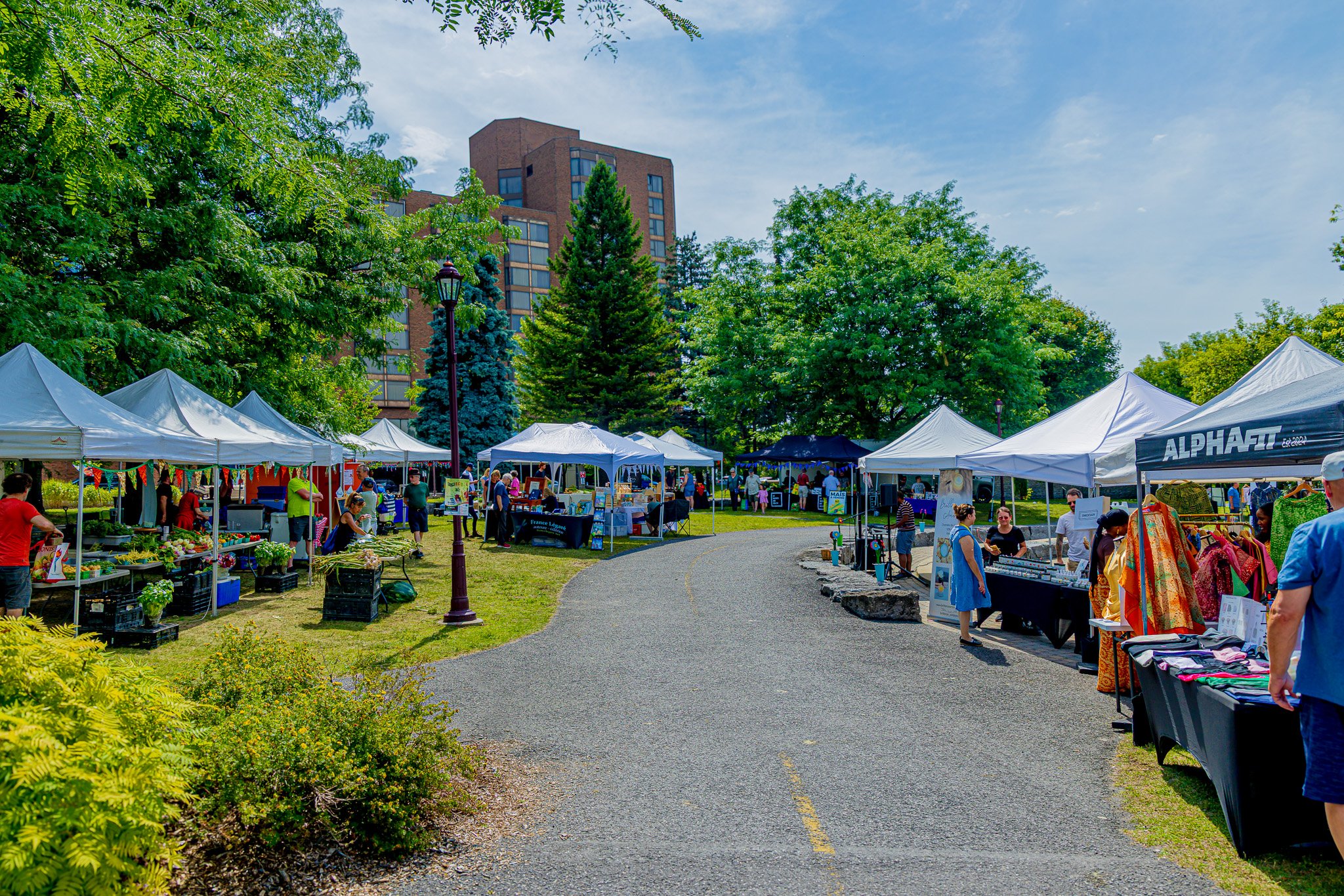 Outdoor market with white tents, vendors selling various goods, and people browsing on a paved path with green trees and a tall building in the background on a sunny day.