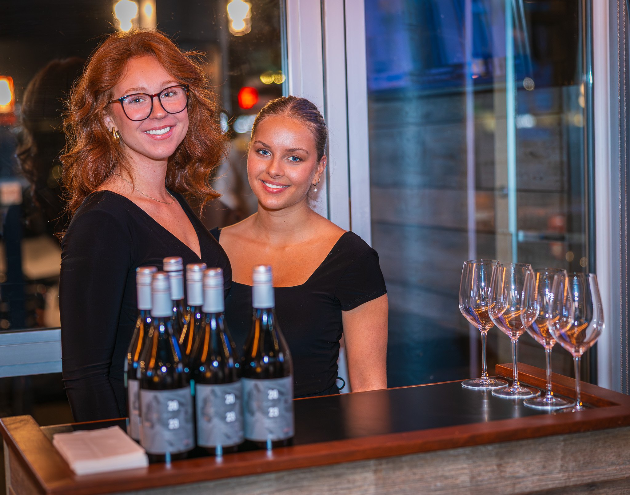 Two women behind a bar counter with bottles of wine and empty glasses, smiling at the camera.