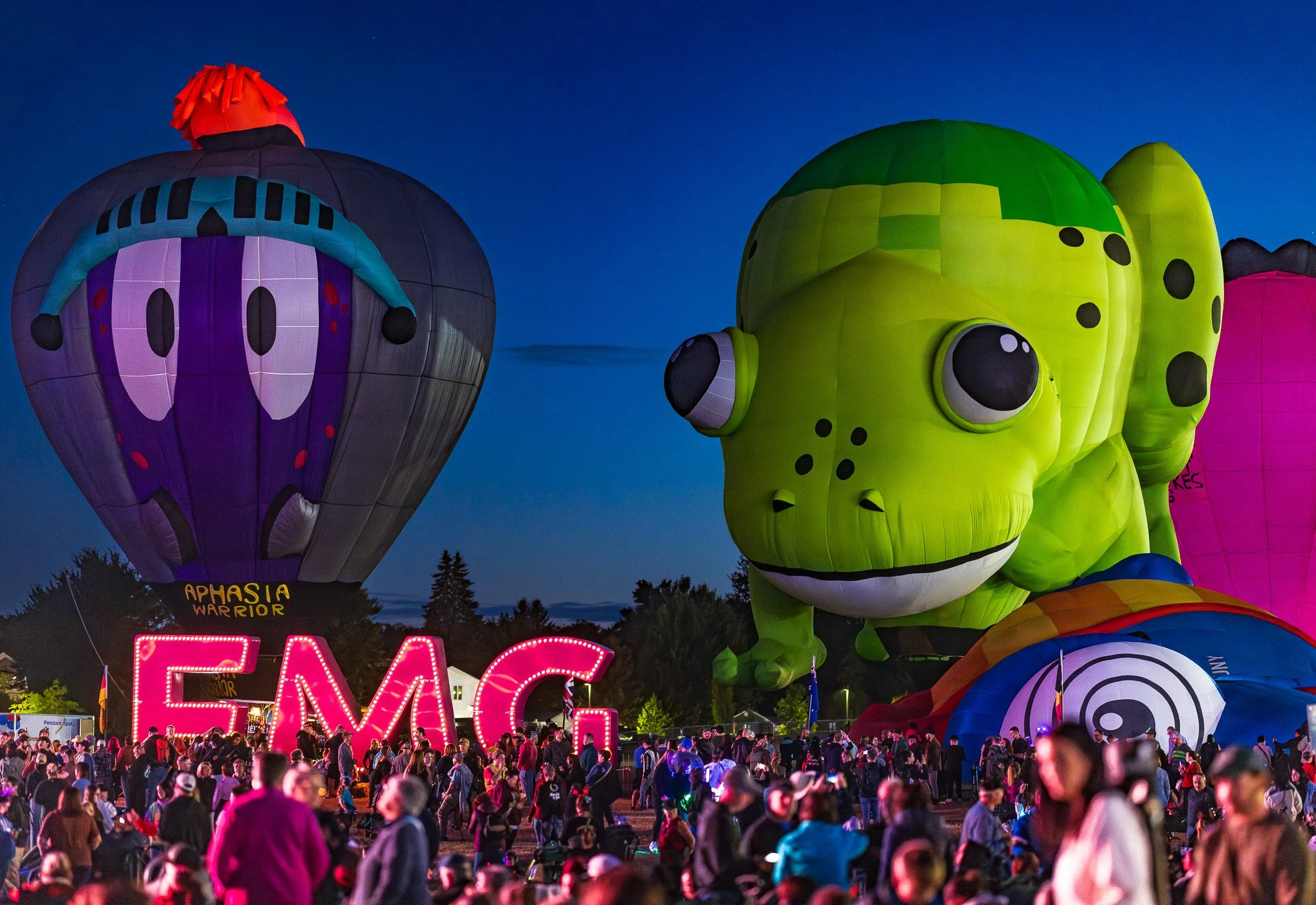 Colorful hot air balloons shaped like a butterfly, a dinosaur, and other figures, with a large crowd of people at a night festival.