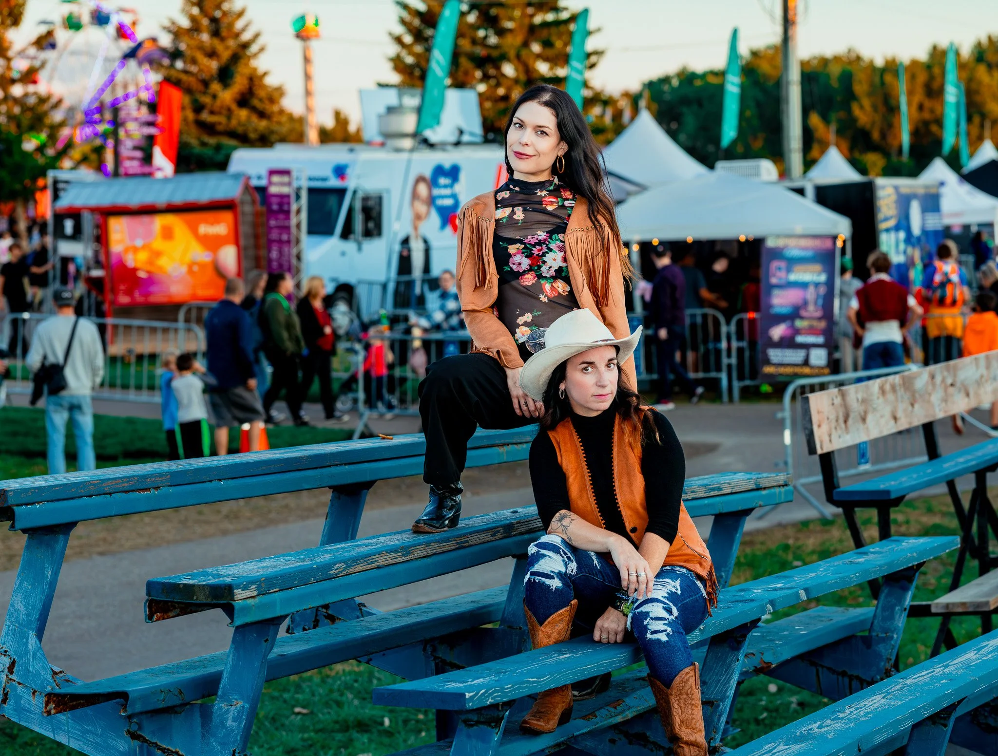 Two women, one seated and one standing, at an outdoor fair with amusement rides and tents in the background. The seated woman wears a white cowboy hat, orange vest, and ripped jeans. The standing woman wears a floral top, fringed jacket, and dark pan