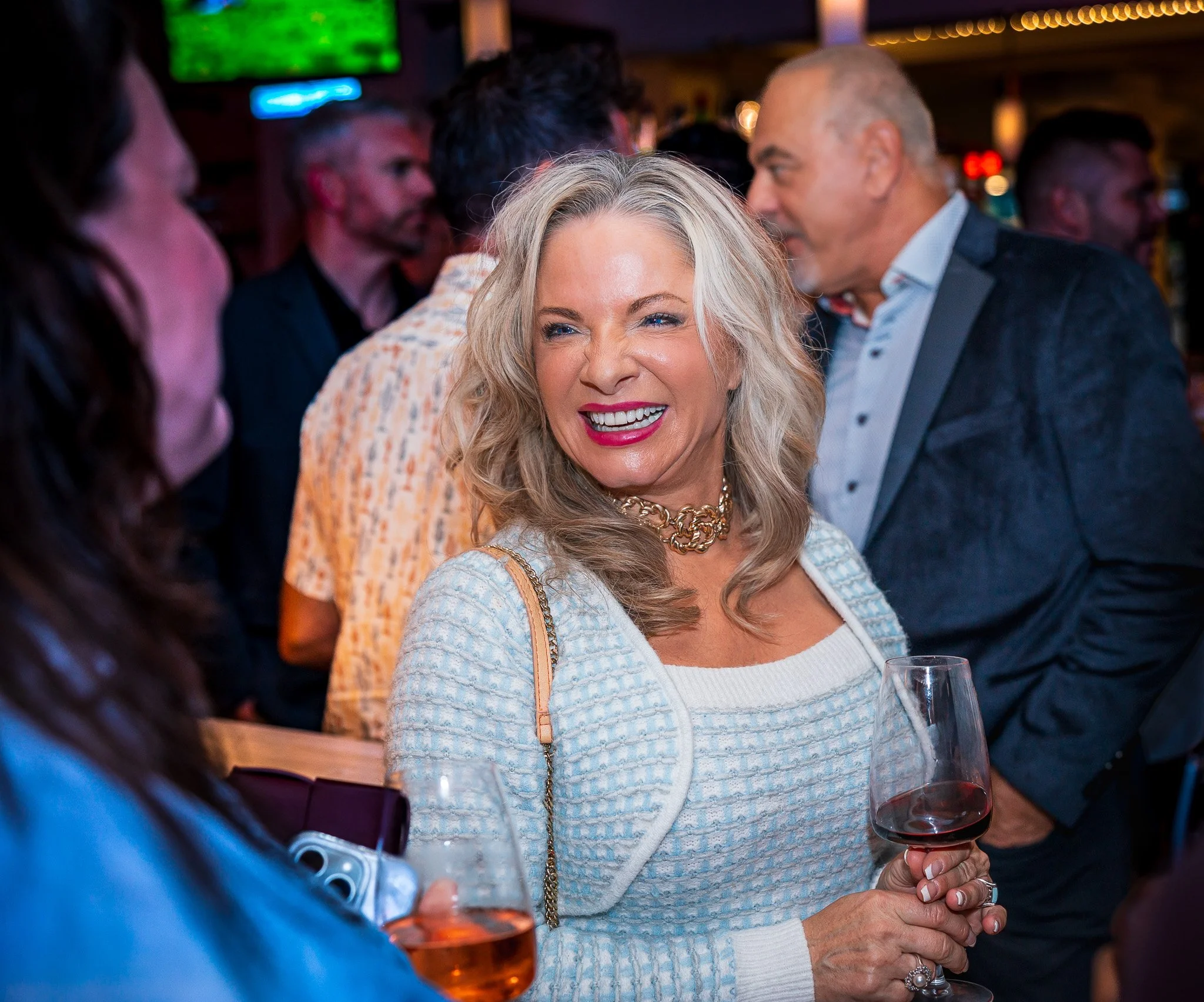 A smiling woman with blonde hair holding a glass of red wine at a social gathering or party, surrounded by other people in conversation in a dimly lit indoor setting.