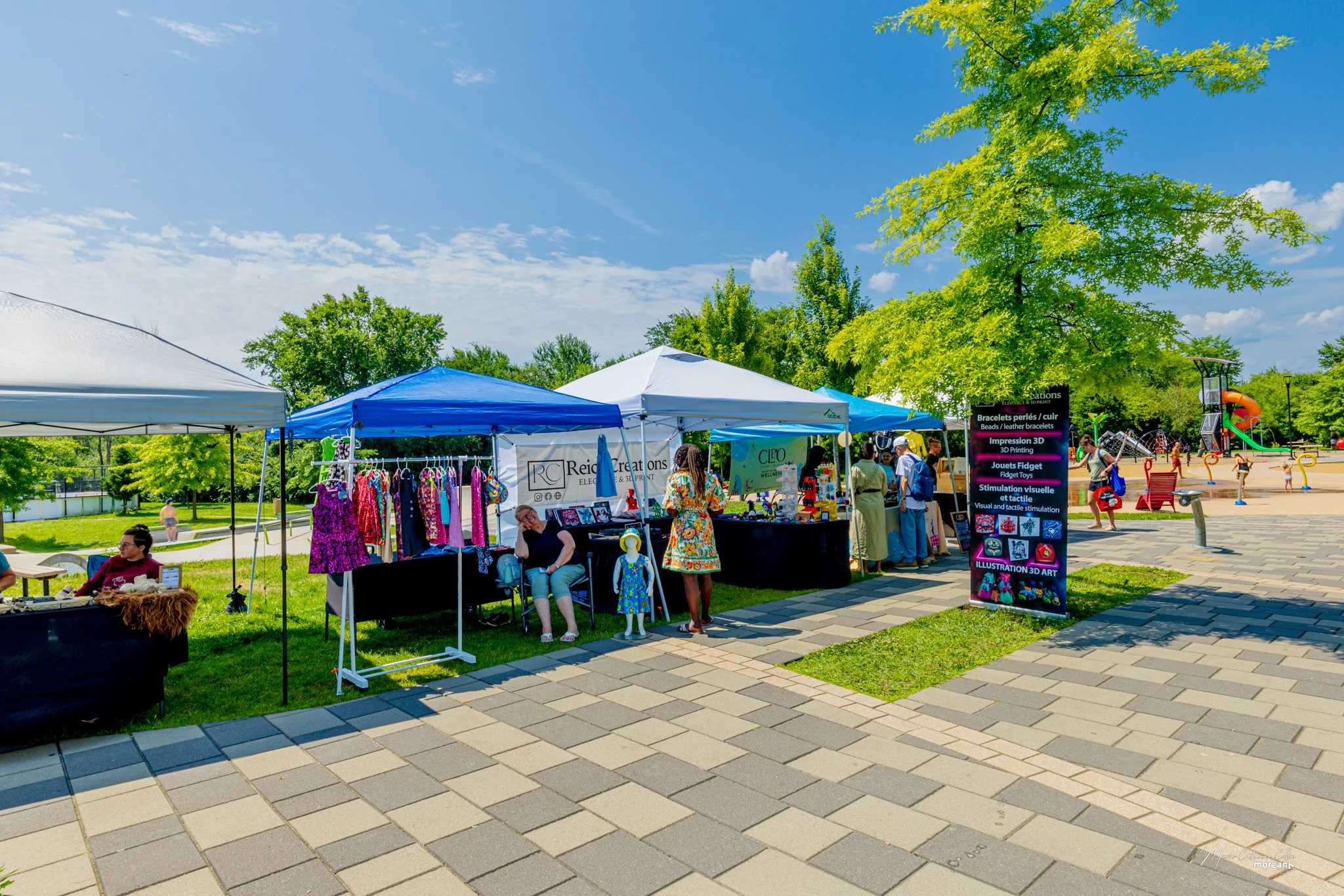 Outdoor market with multiple tents selling clothing and jewelry, people browsing, and a playground with children playing in the background on a sunny day.