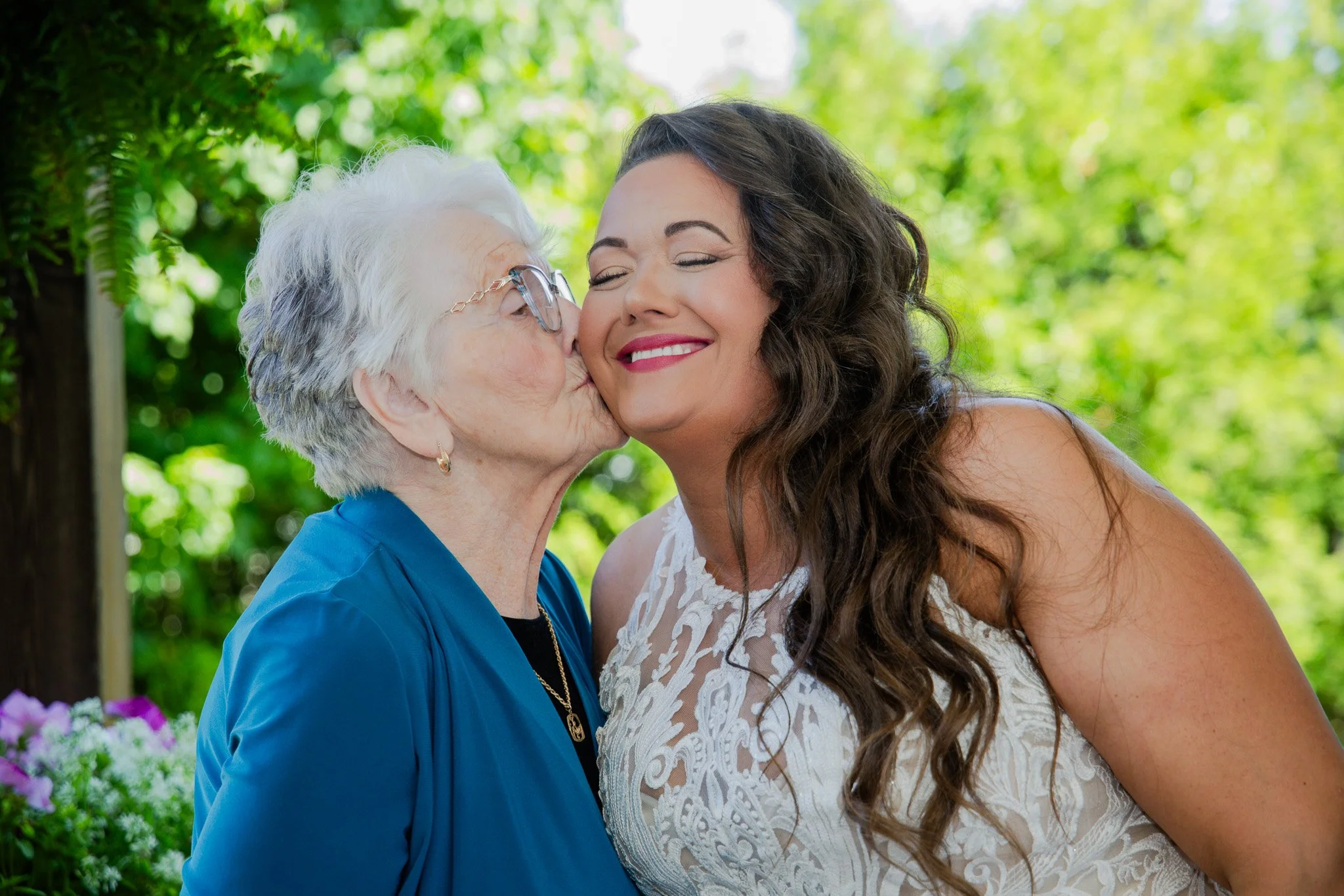 An elderly woman with white hair and glasses kisses a smiling woman with long, dark, curly hair on the cheek outdoors with green trees and plants in the background.