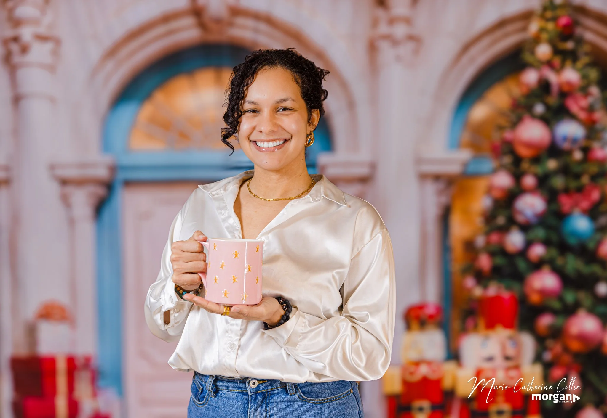 A woman with short curly dark hair smiling and holding a pink mug with gold star designs in front of a decorated Christmas tree and festive background.
