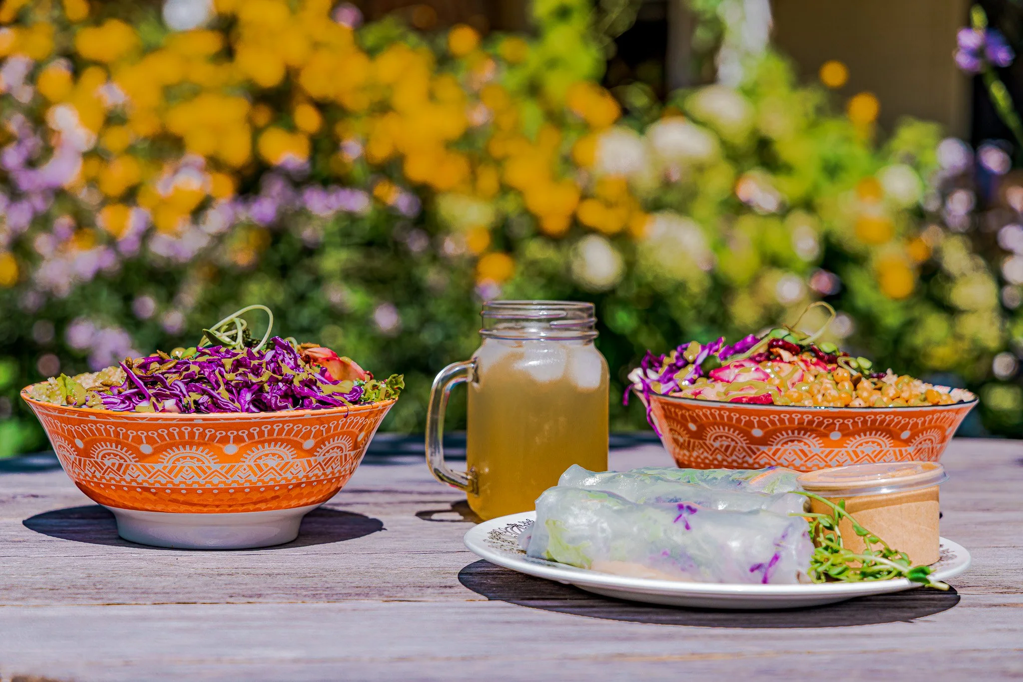 Colorful salads in orange patterned bowls, a small jar of dressing, and spring rolls on a white plate, with a glass jar of drink in the center, on a wooden table outdoors with blurred yellow and purple flowers in the background.