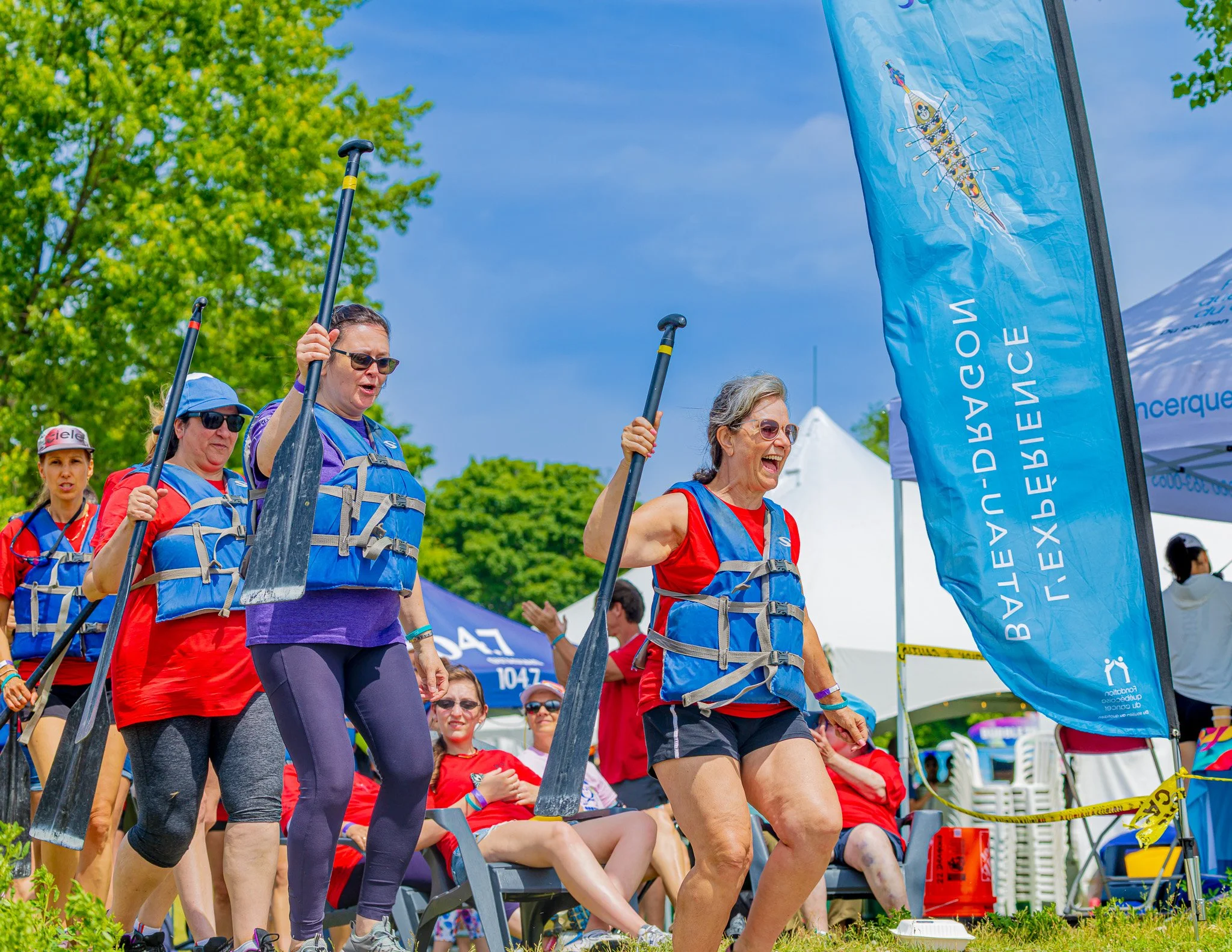 Women wearing life jackets and holding paddles at a water sports event, with a blue banner and tents in the background.