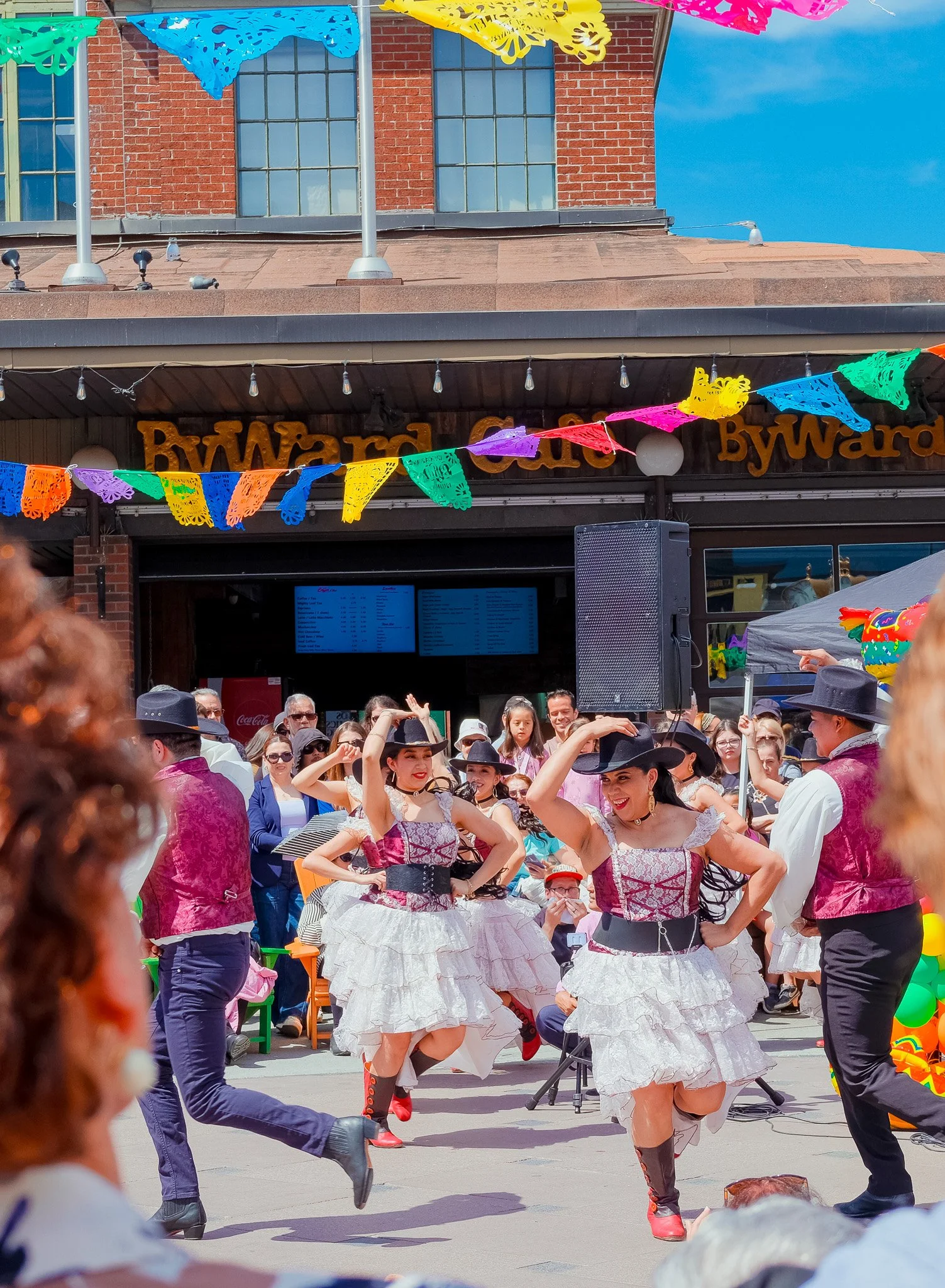 People celebrating at a festival with dancers dressed in traditional costumes, colorful paper banners hanging overhead, and a crowd of onlookers watching the performance.
