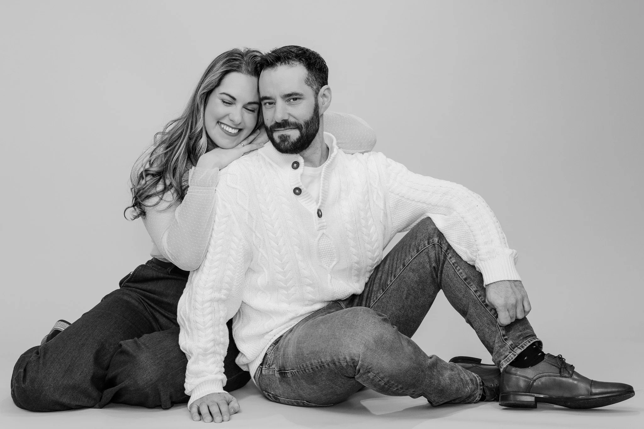 Black and white photo of a smiling woman hugging a seated man with a beard. The woman has long wavy hair, and the man is wearing a white knit sweater and dark jeans. They are sitting on the floor against a plain backdrop.